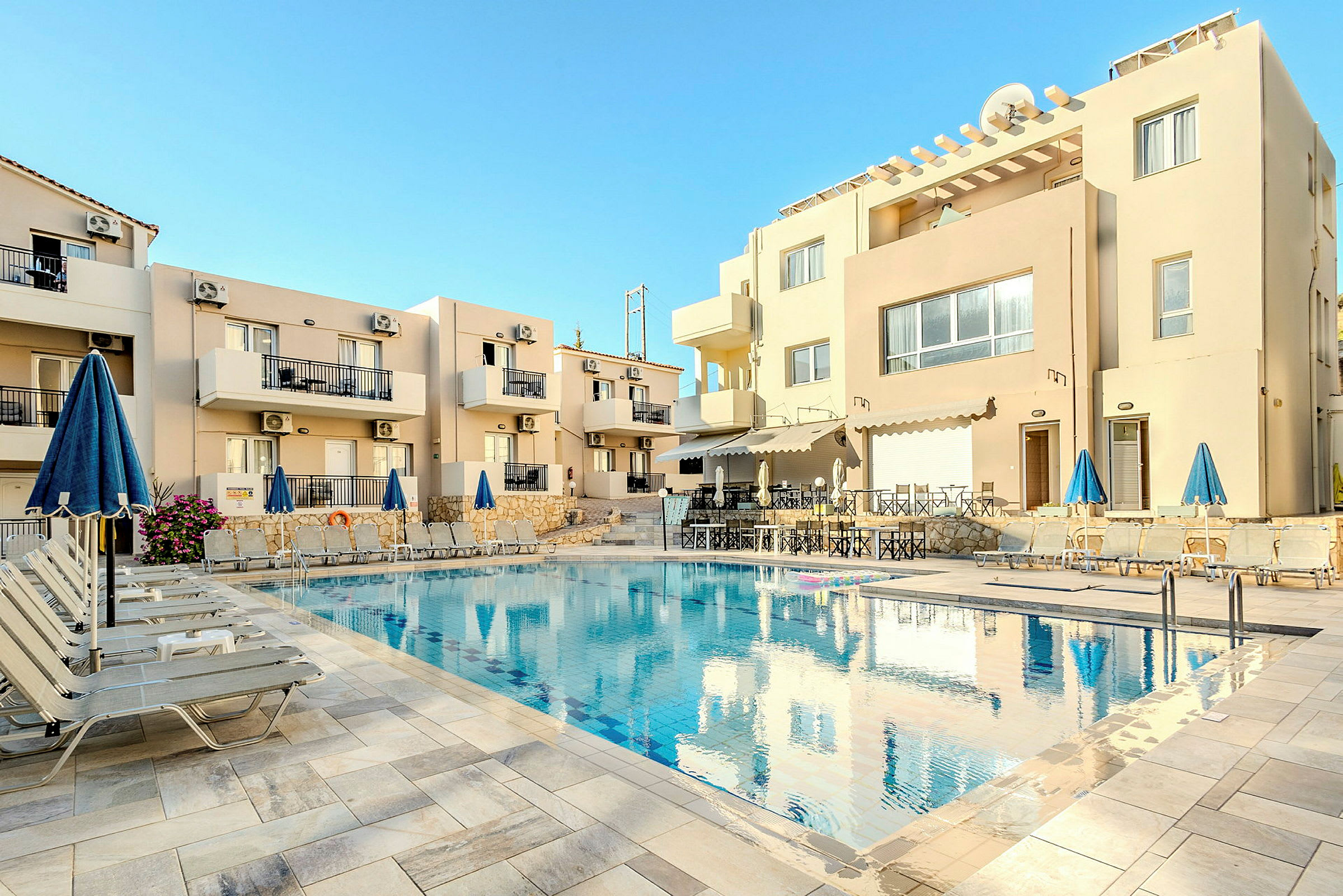 Udendørs swimmingpool omgivet af liggestole og parasoller foran en beige beboelsesbygning. (AI-genereret tekst)