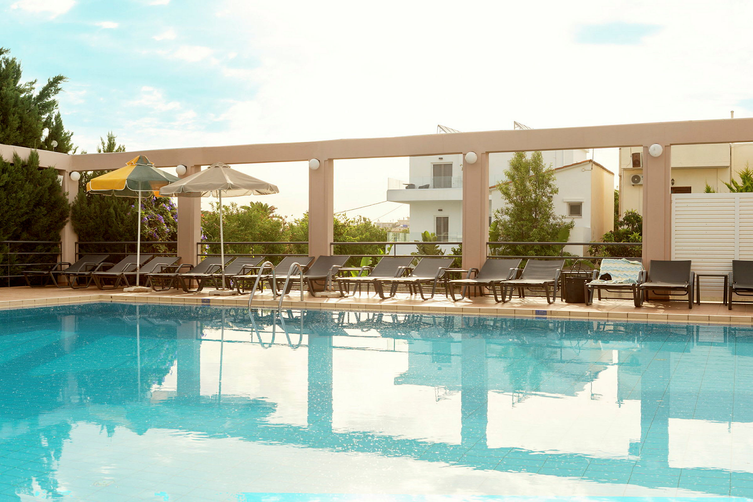 En swimmingpool med liggestole og parasoller rundt kanten, omgivet af vegetation og bygninger. (AI-genereret tekst)