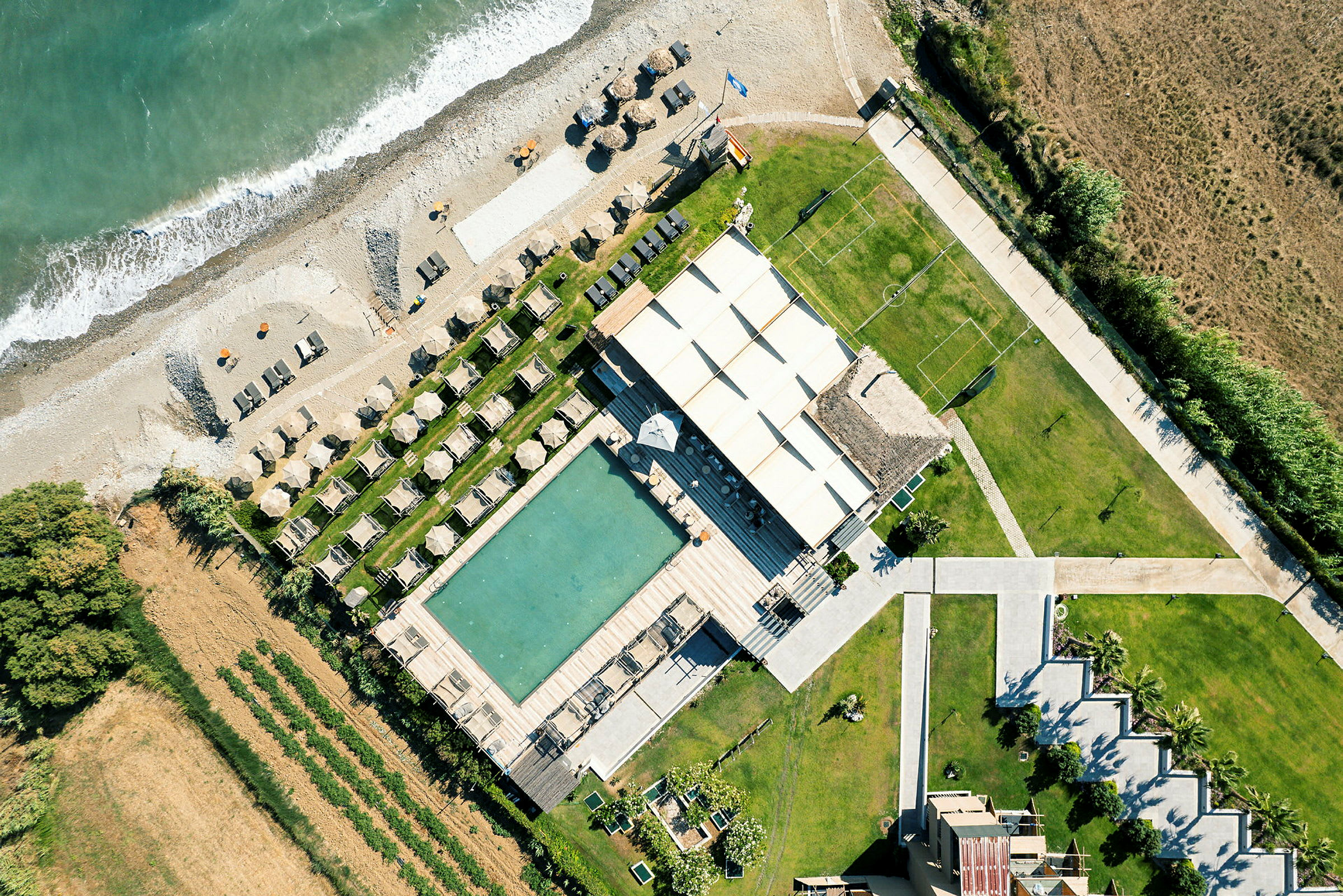 Luftfoto af et strandnært resort med liggestole, parasoller, pool og en frodig omgivende natur. (AI-genereret tekst)