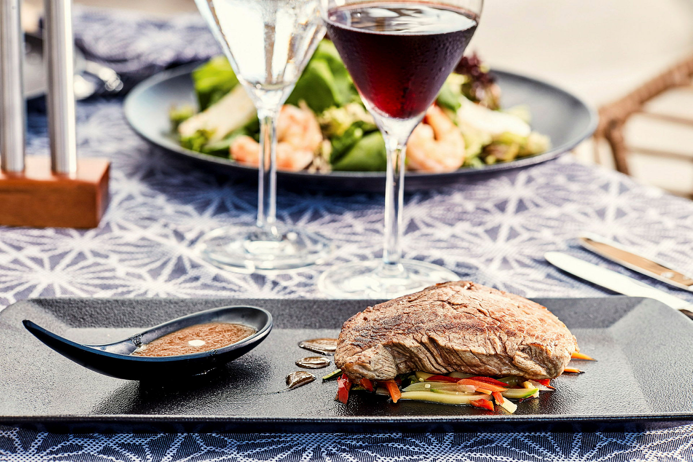 En tallerken med kød på grøntsager og en lille sauce skål, med vinglas og salat i baggrunden. (AI-genereret tekst)