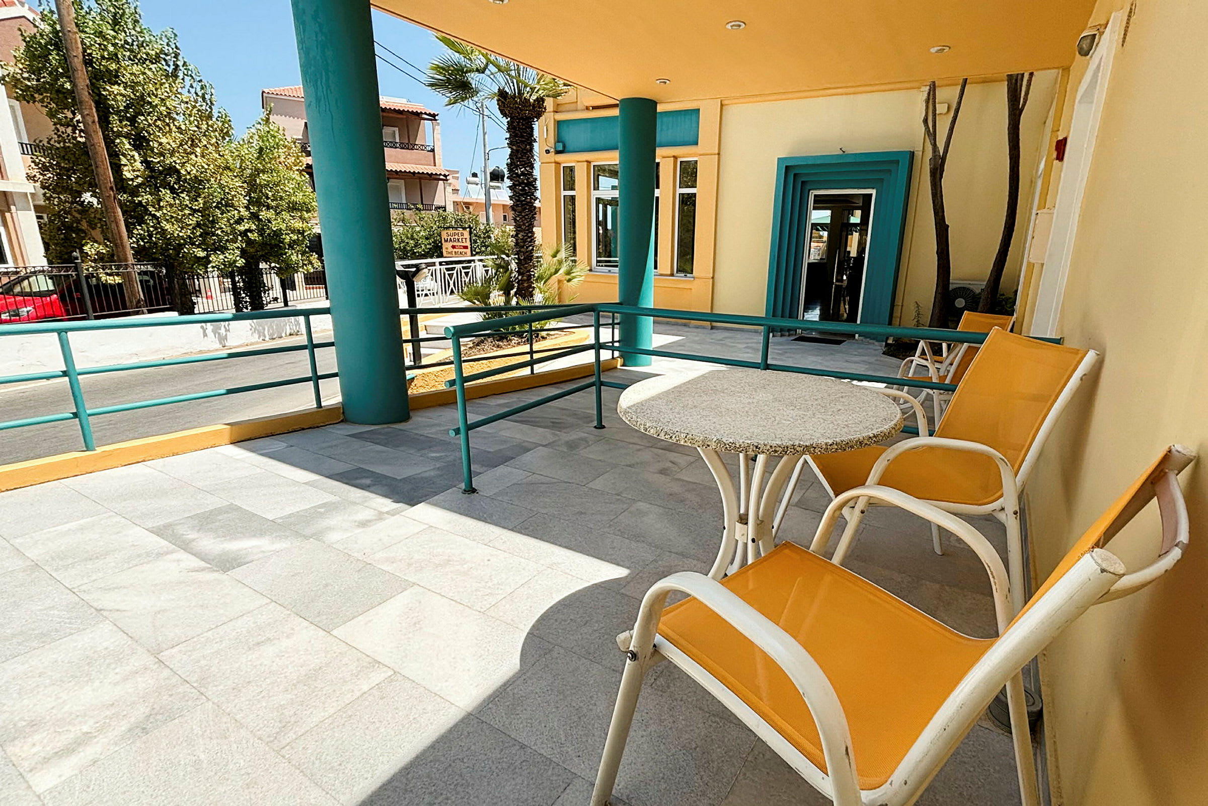 En terrasse med bord og stole, omgivet af farverige bygninger og træer. Der ses en vej i baggrunden. (AI-genereret tekst)