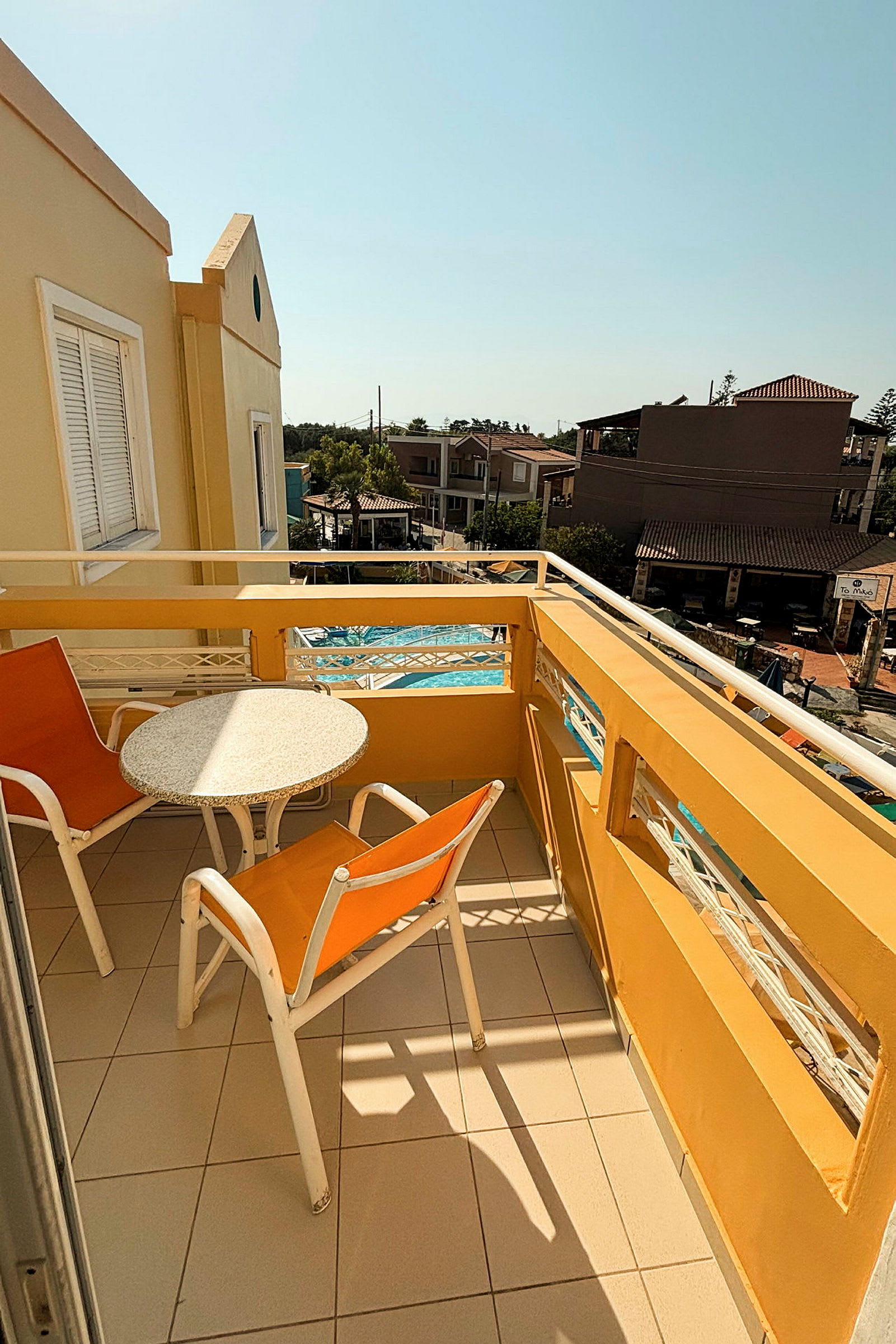 En gul balkon med bord og to stole, udsigt over bygninger og pool. (AI-genereret tekst)