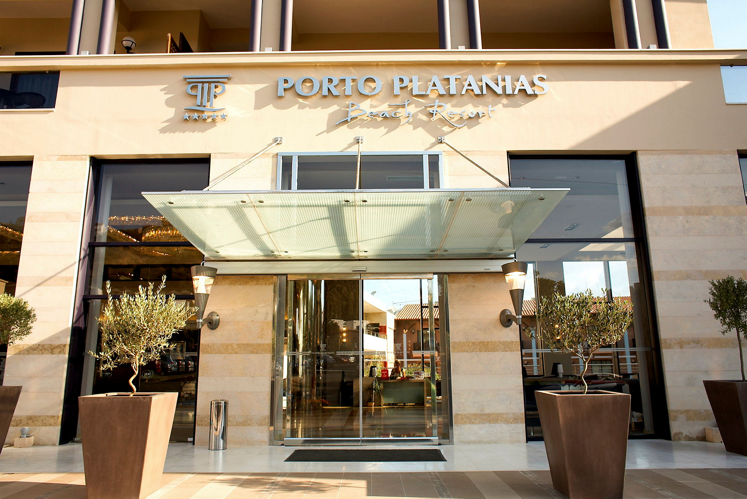 Indgangen til Porto Platanias Beach Resort, med glasdøre og plantede oliventræer foran. (AI-genereret tekst)