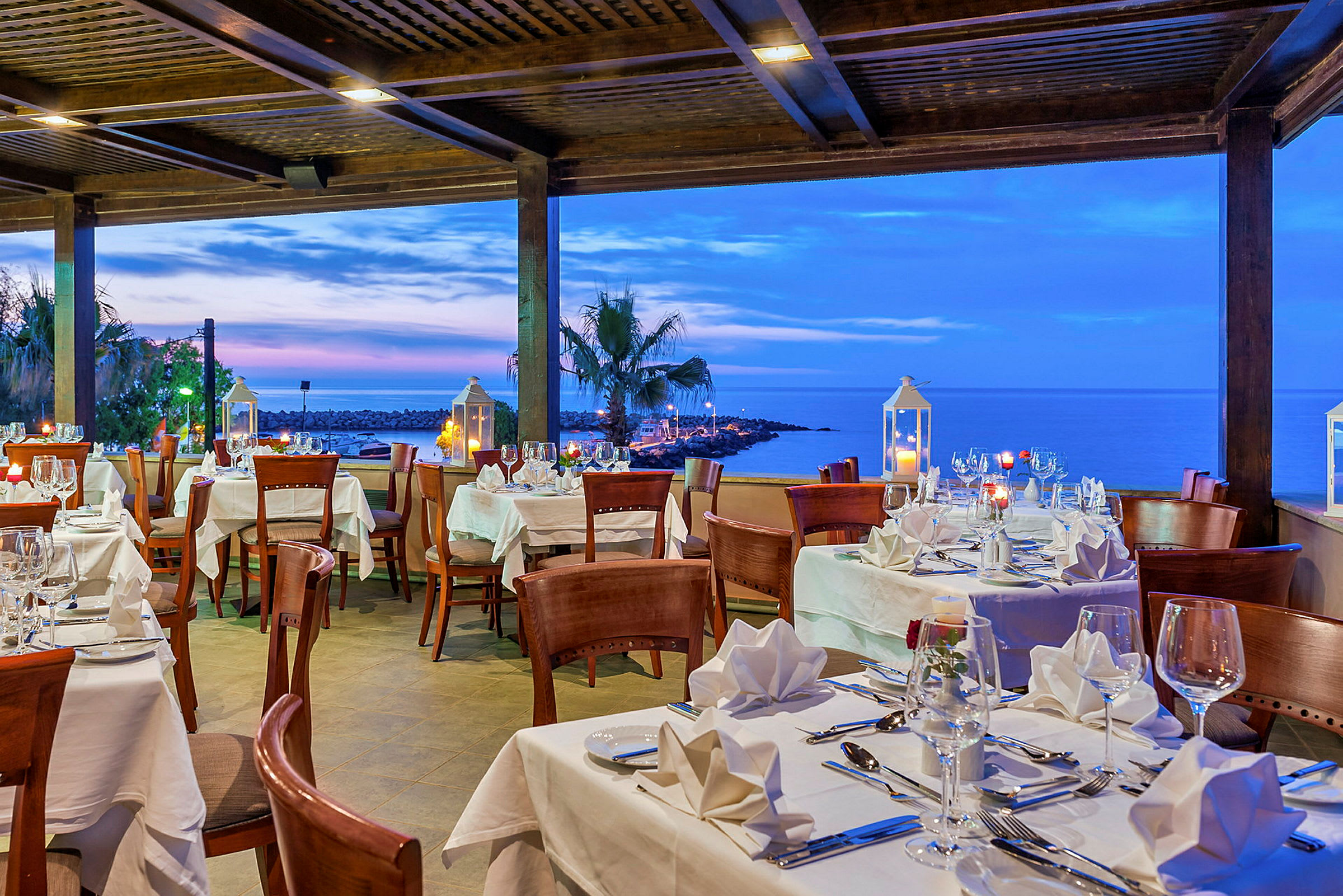 En restaurant med dækkede borde med hvide duge, med udsigt over havet og solnedgangen. (AI-genereret tekst)