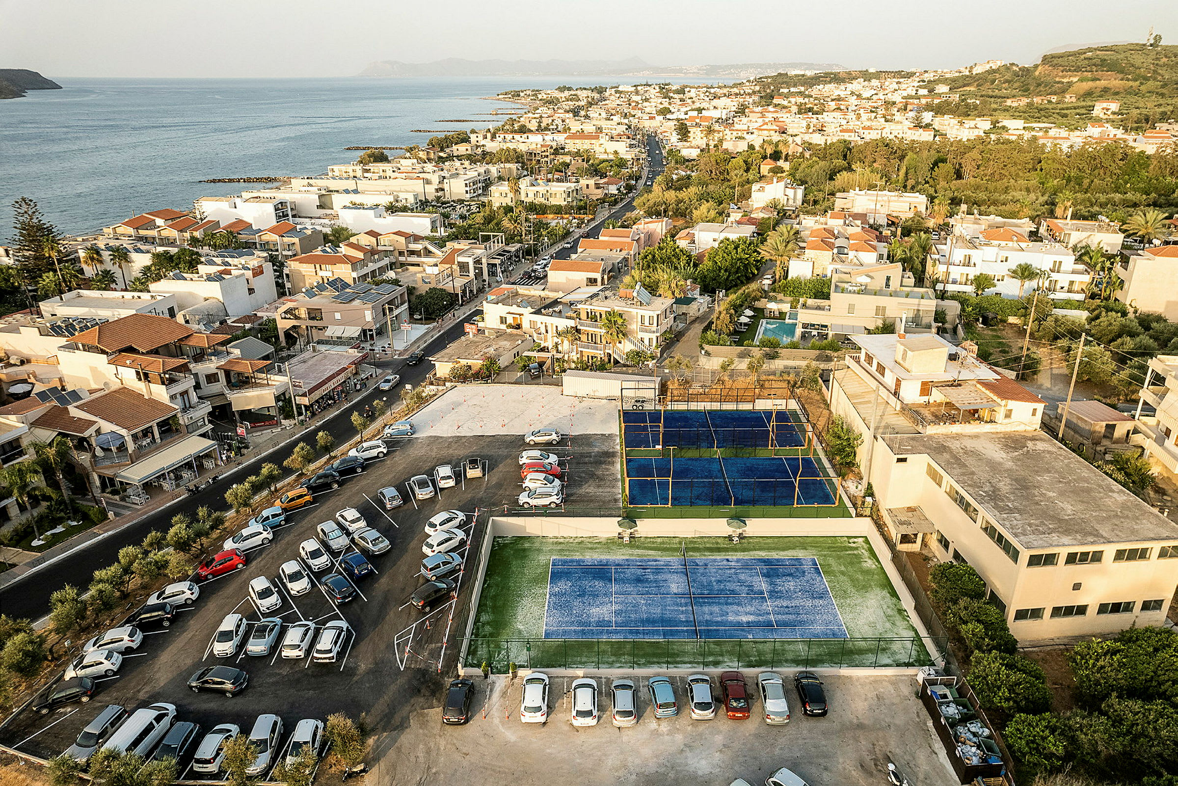 Luftfoto af et kystnært boligområde med tennisbaner og parkeringsplads. (AI-genereret tekst)