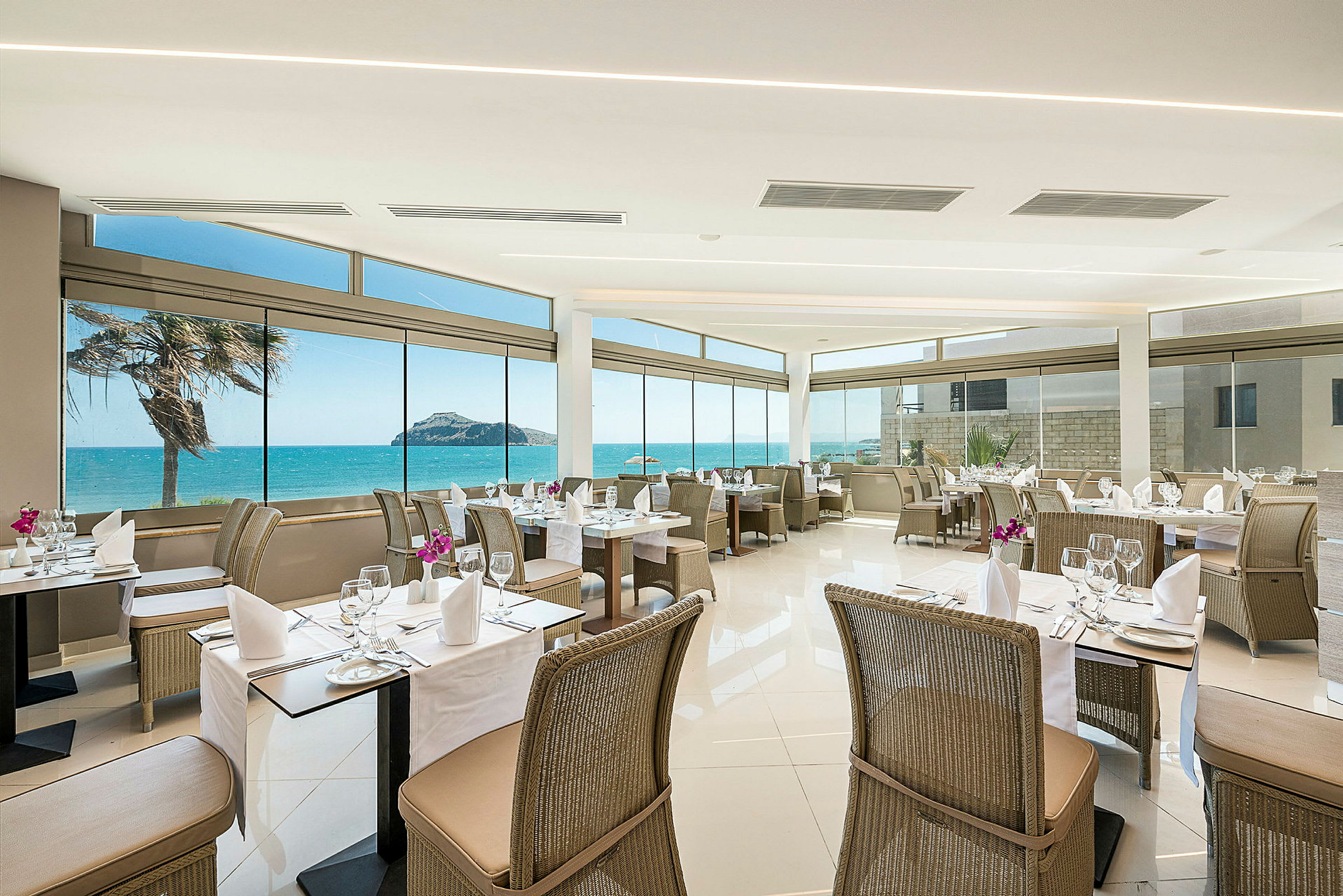 Restaurant med udsigt til havet, dækkede borde og rottingstole. (AI-genereret tekst)