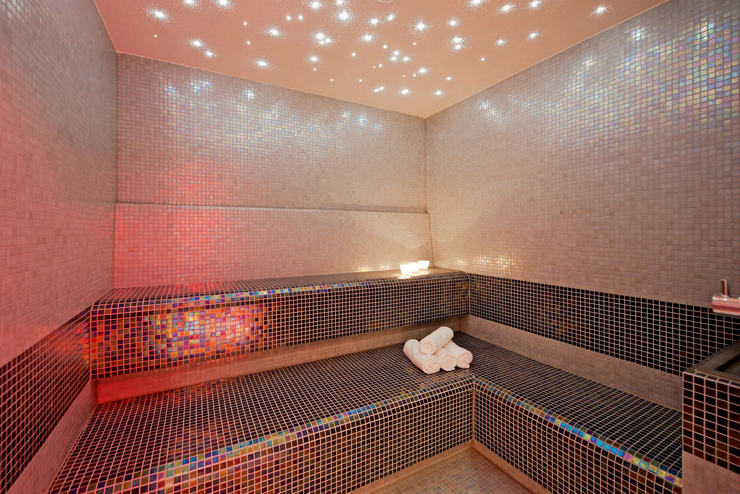 Rummet er et mosaikflisebelagt spa med en bænk og håndklæder. (AI-genereret tekst)