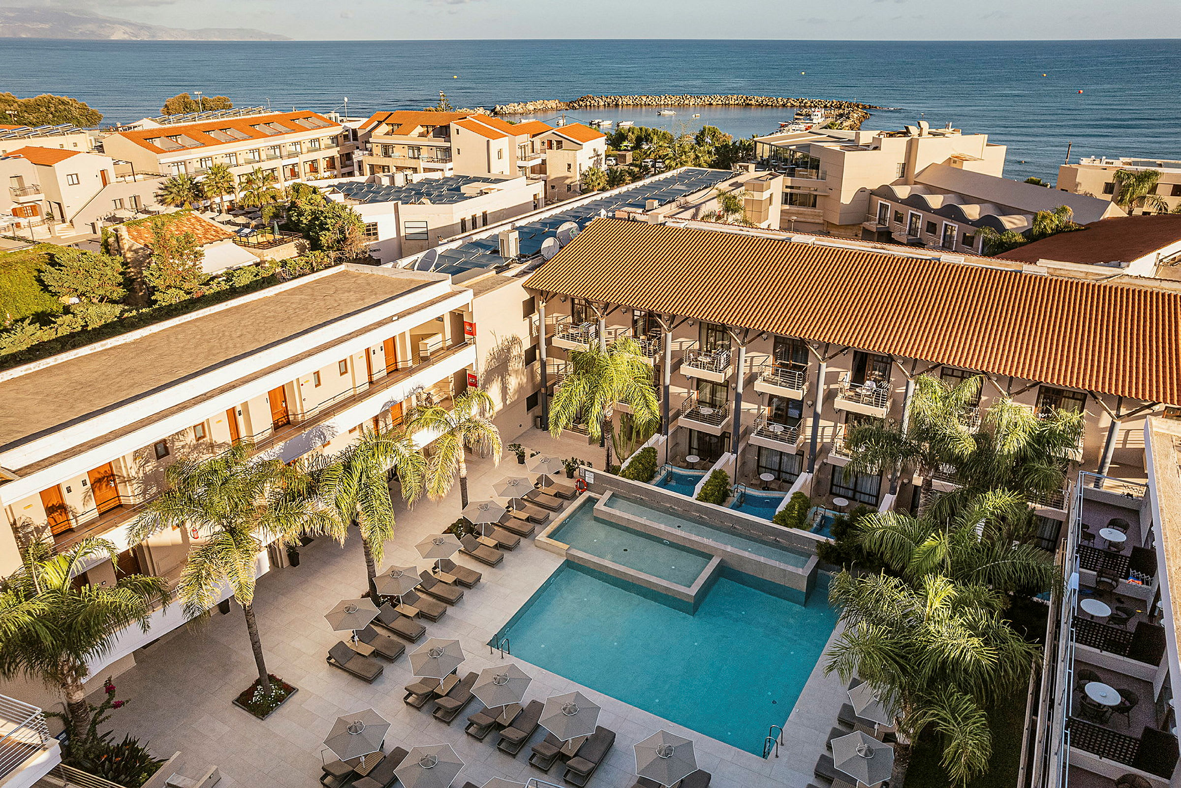 Hotelområde med pool, liggestole og bygninger, samt havet i baggrunden. (AI-genereret tekst)