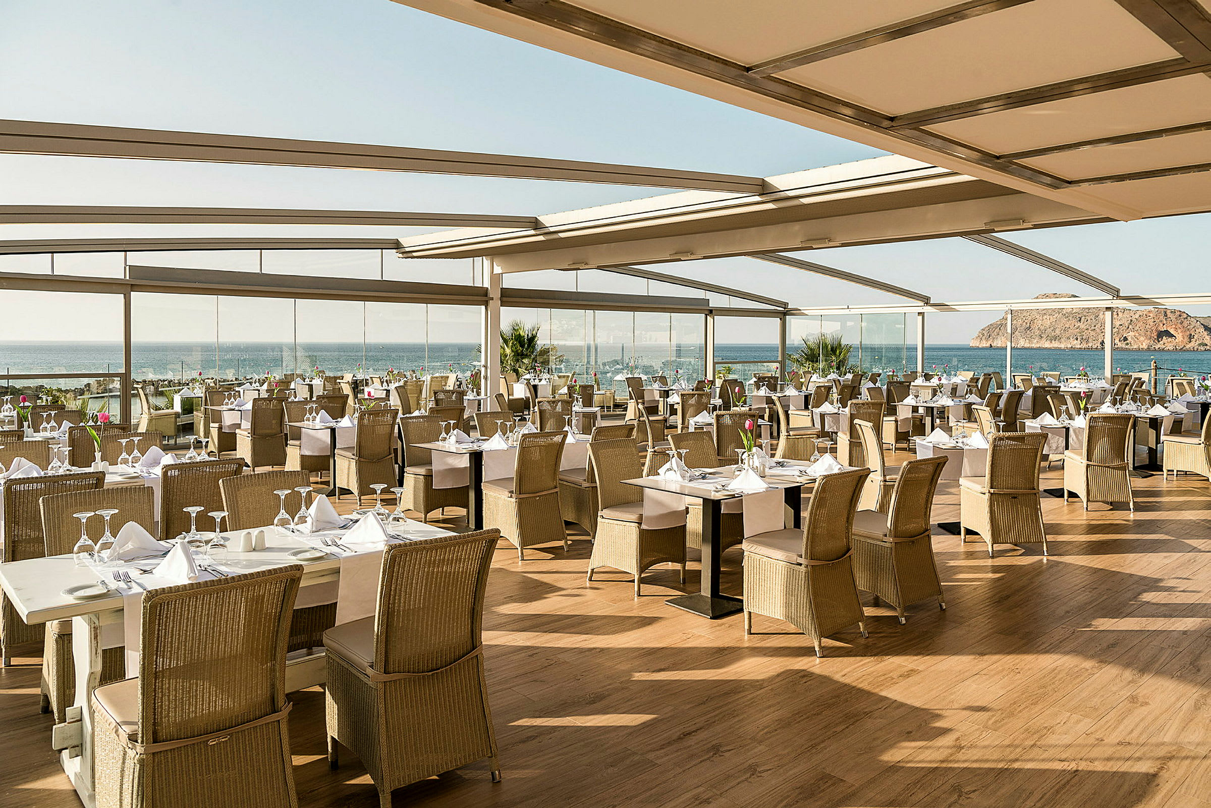 En restaurant med dækkede borde og udsigt over havet, omgivet af store glasvægge og tag. (AI-genereret tekst)