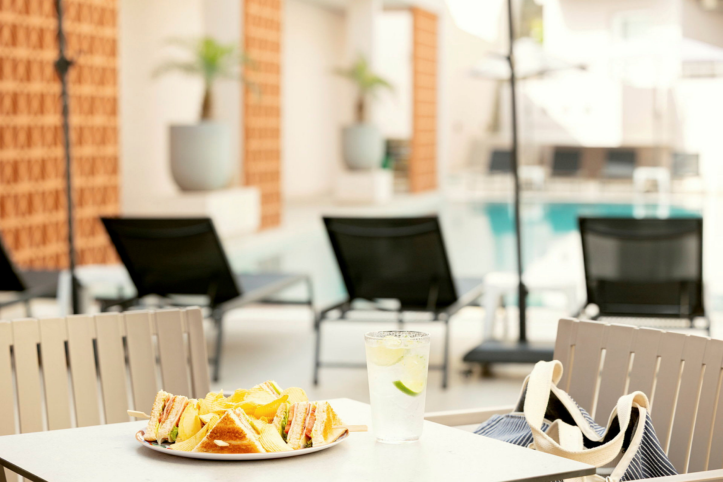 En tallerken med sandwiches og chips samt et glas vand med lime står på et bord ved en pool med liggestole i baggrunden. (AI-genereret tekst)