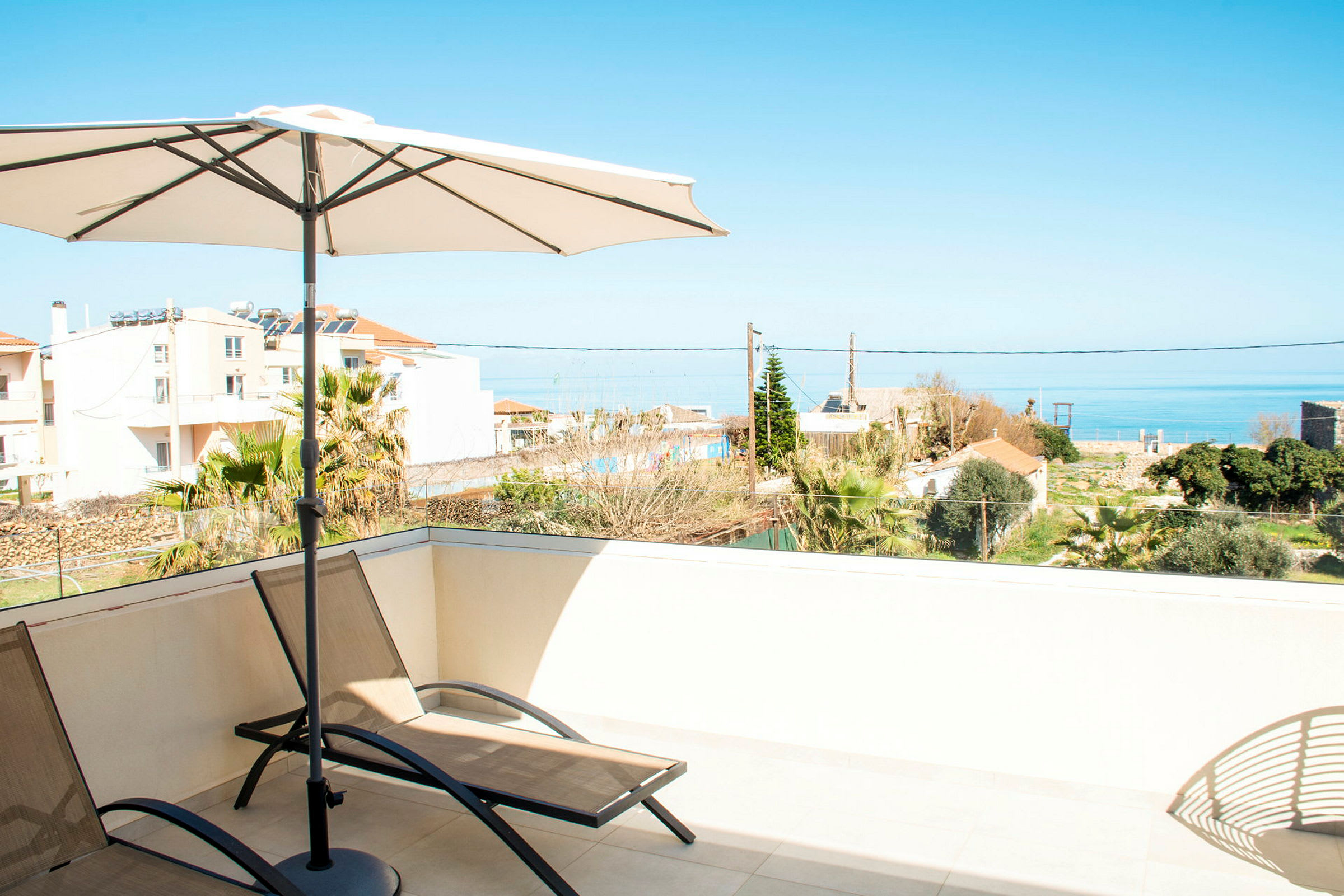 Liggestole og parasol på en terrasse med udsigt over havet og omkringliggende bygninger. (AI-genereret tekst)
