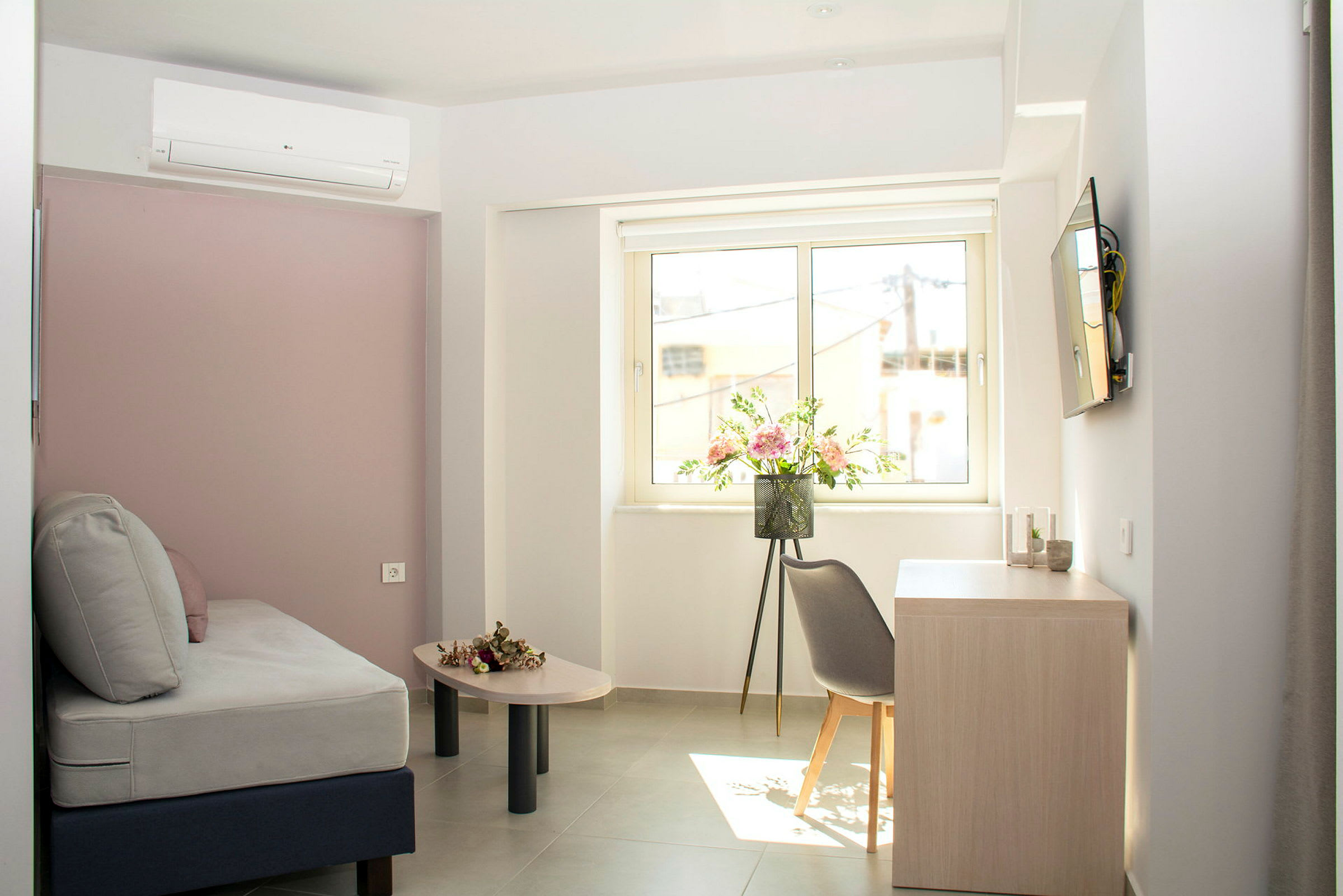 Et lyst hotelværelse med grå sofa, blomster på bordet og vinduesudsigt. (AI-genereret tekst)