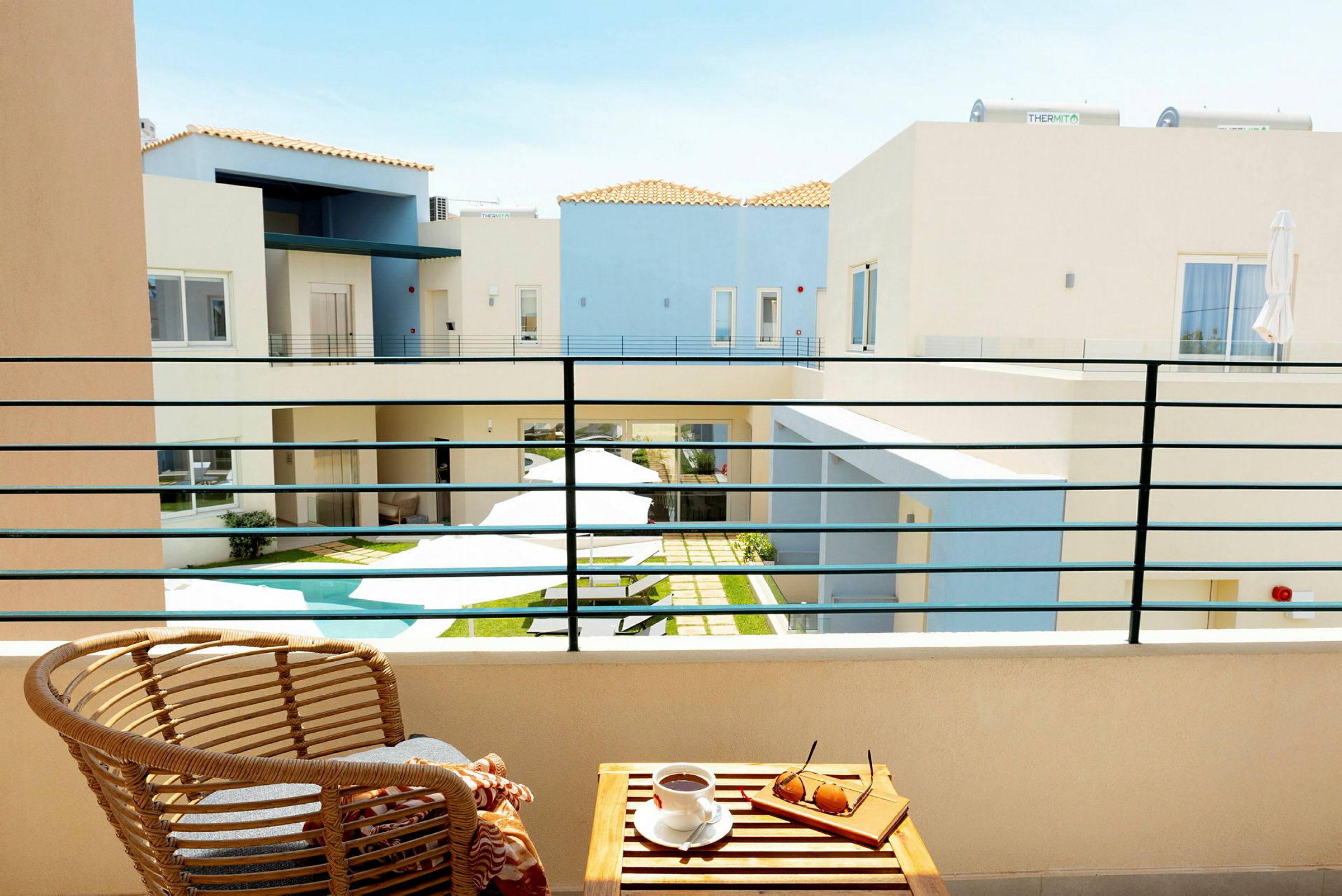 Udsigt fra en balkon til en gårdhave med en lille pool omgivet af lyse bygninger. På balkonen er der en stol og et bord med solbriller og en kaffekop. (AI-genereret tekst)