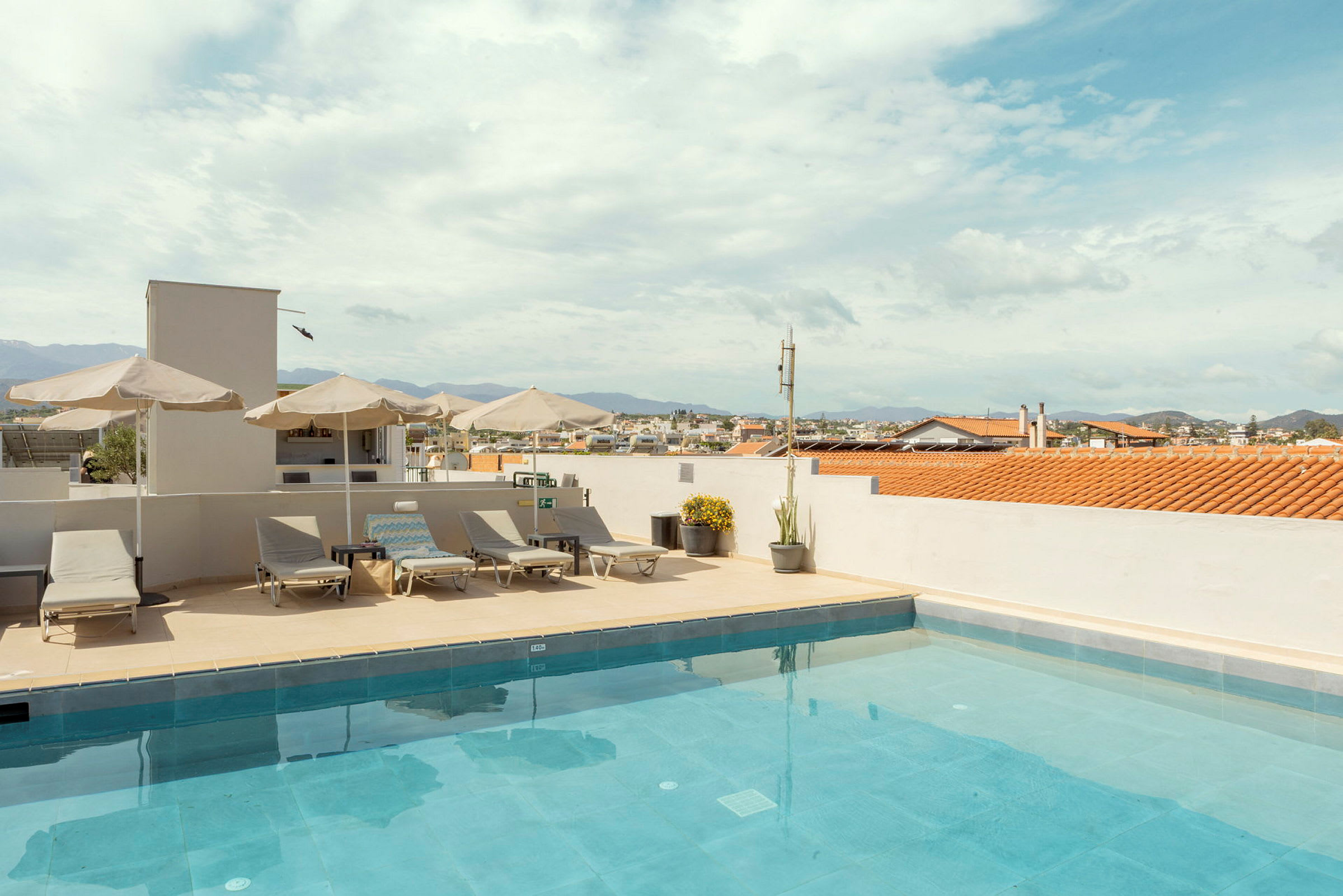 En tagterrasse med en swimmingpool, liggestole og parasoller under en blå himmel med udsigt over byen. (AI-genereret tekst)