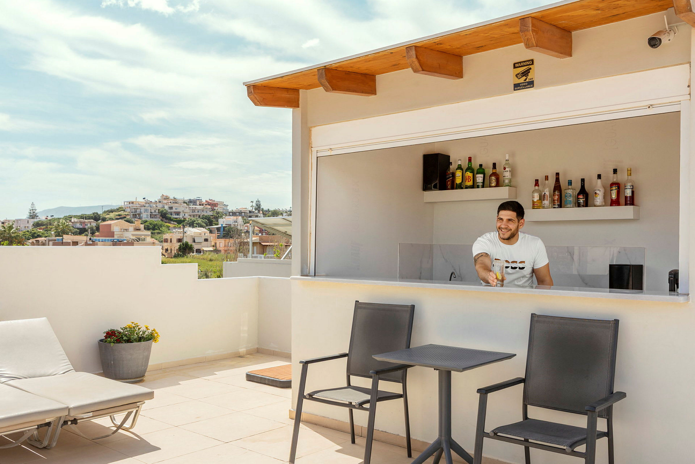 En terrasse med en bar, hvor en person står bag disken. Foran baren er der borde med stole og en liggestol. (AI-genereret tekst)