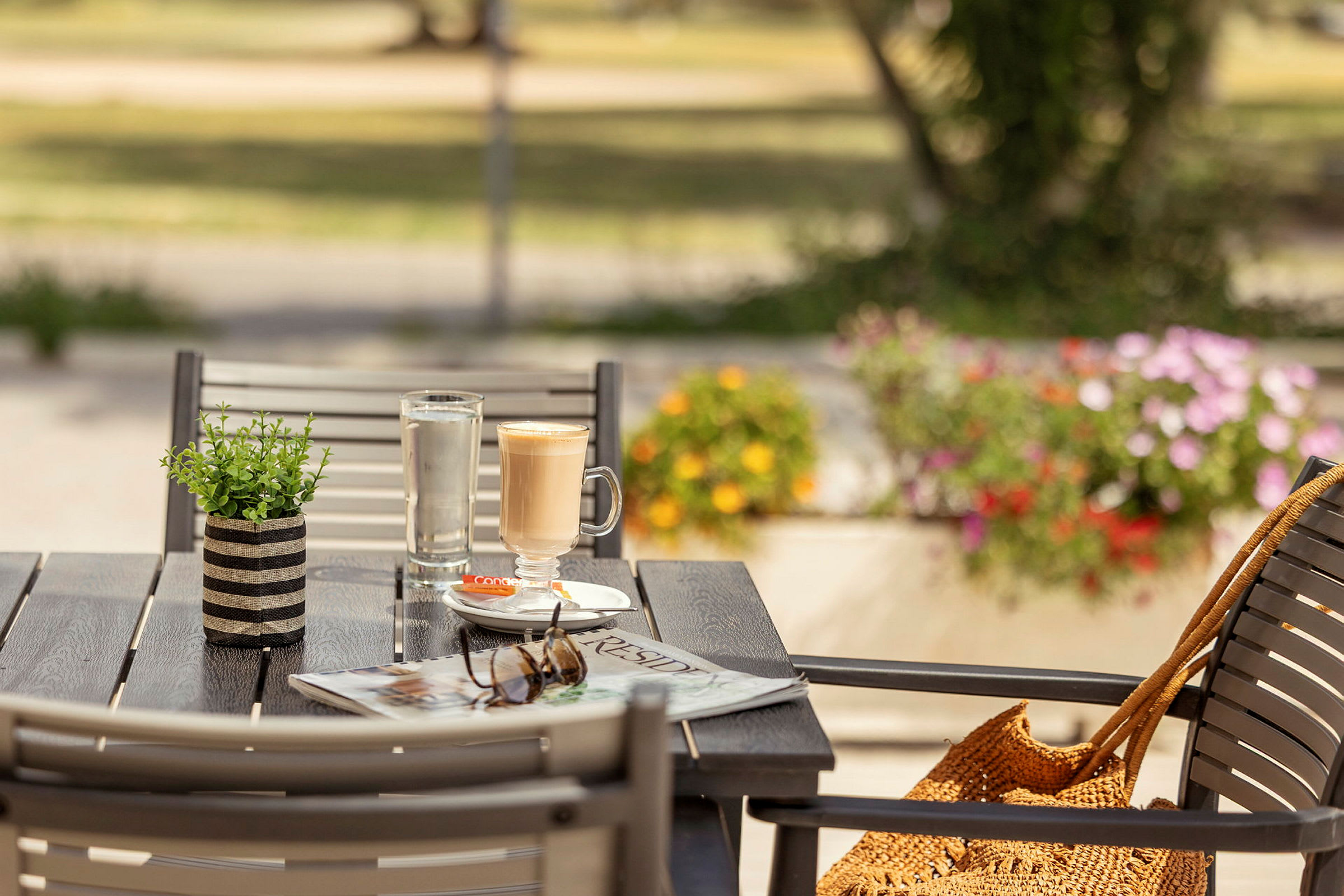 En udendørs servering med et bord, der har en kaffekop, et glas vand, en avis og solbriller. Baggrunden viser et grønt område og blomster. (AI-genereret tekst)