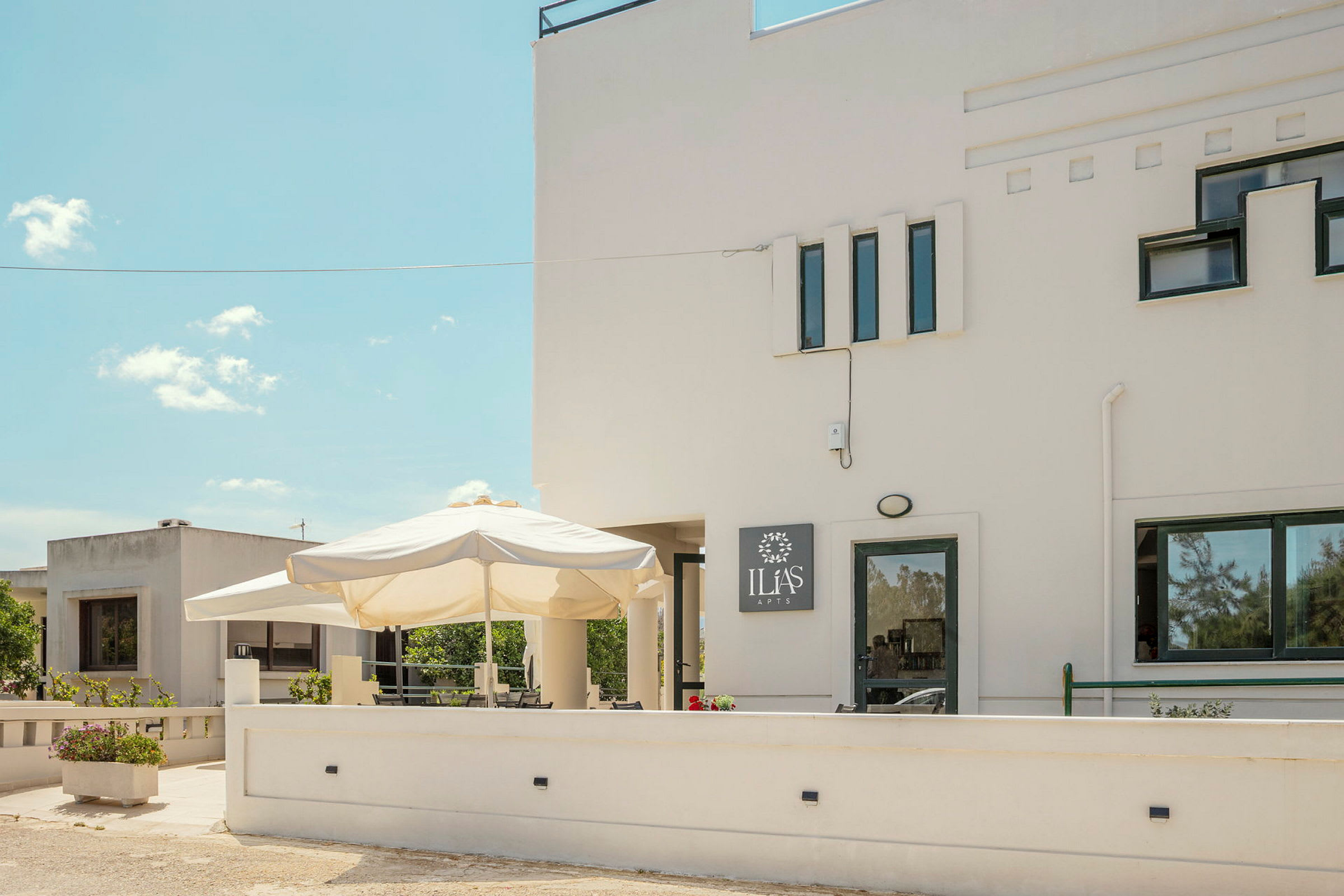 Billede af en hvid bygning med flere vinduer og et skilt, der siger 'Ilias Apts'. Foran bygningen er der en terrasse med en stor parasol og blomsterkasser. (AI-genereret tekst)