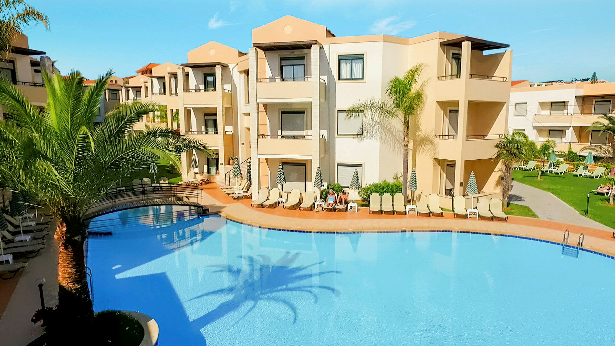 En udendørs pool omgivet af liggestole og palmer ved en beige hotelbygning. (AI-genereret tekst)