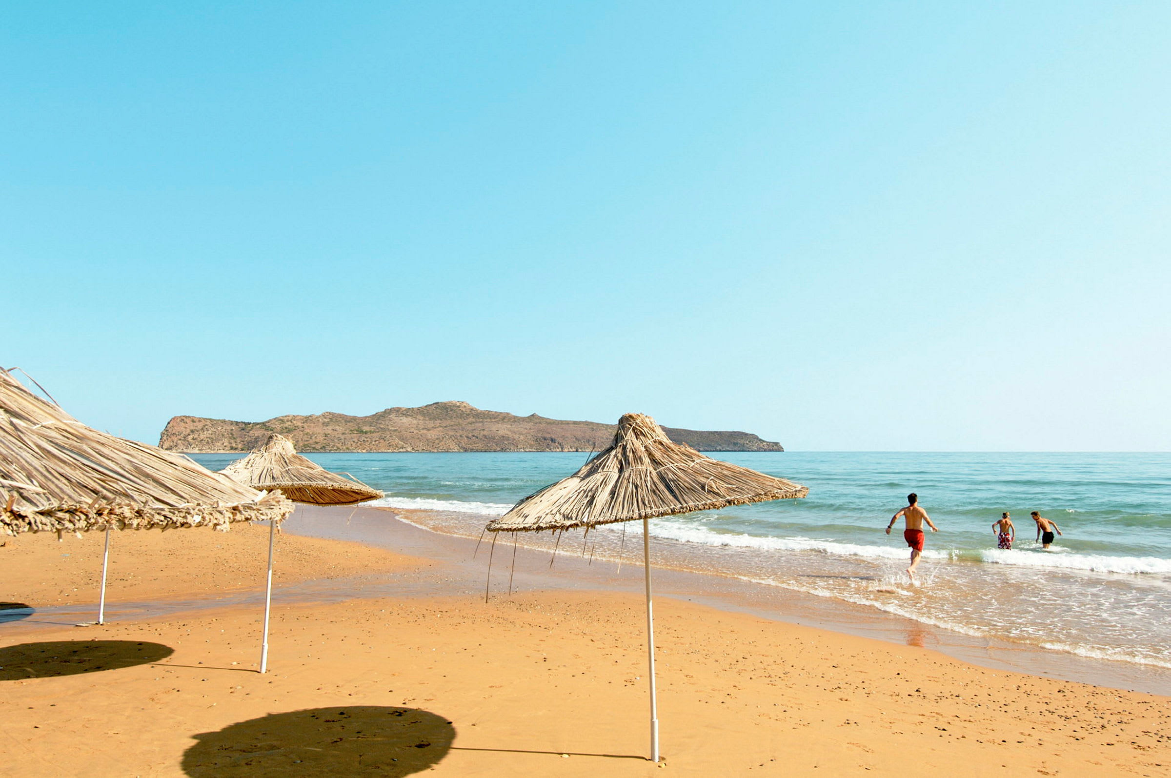 Strand med parasoller af siv og mennesker, der bader i havet. (AI-genereret tekst)