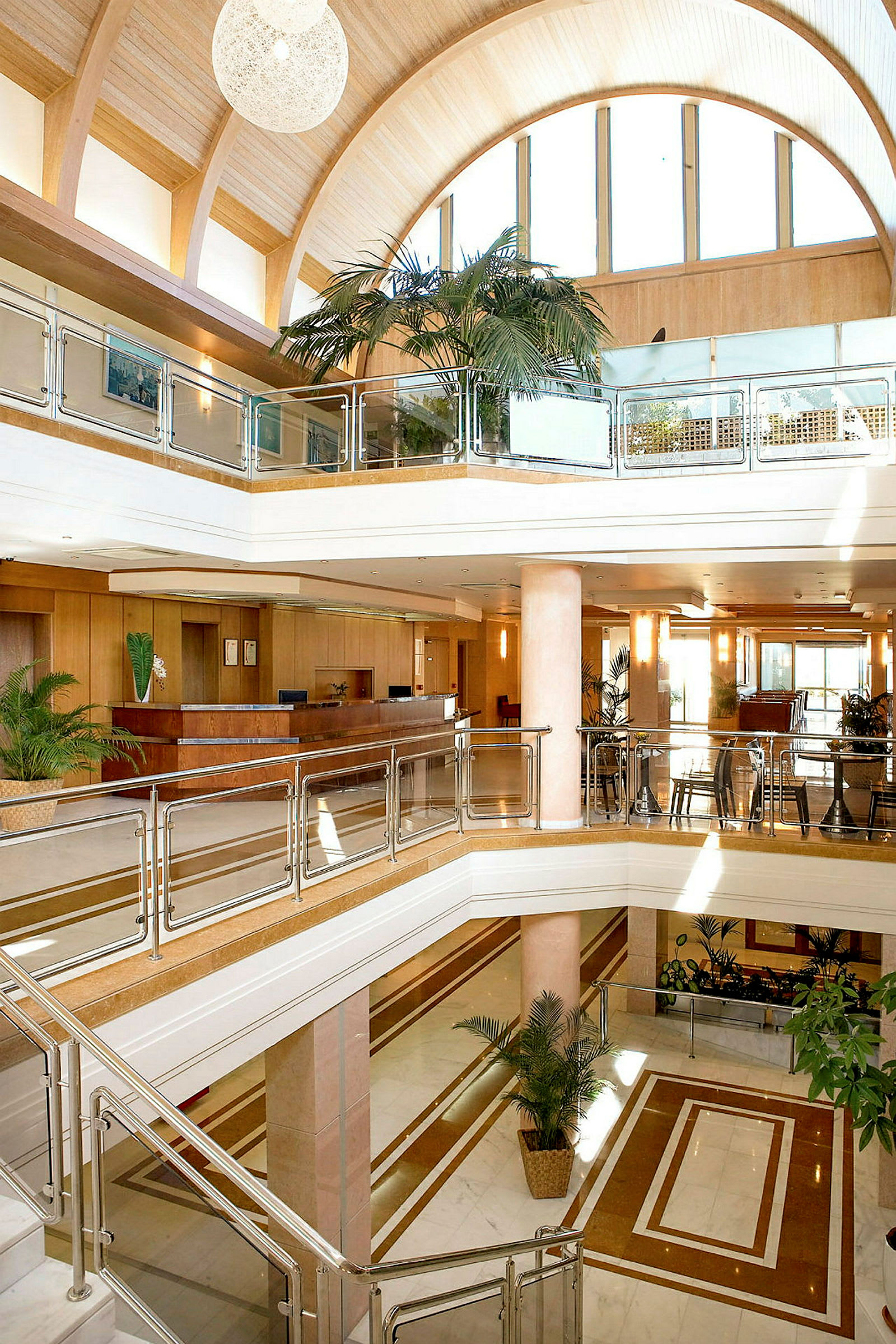 Interiøret i en moderne hotellobby med flere etager. Glasgelændere, trædetaljer og store palmer ses sammen med en reception i baggrunden. (AI-genereret tekst)