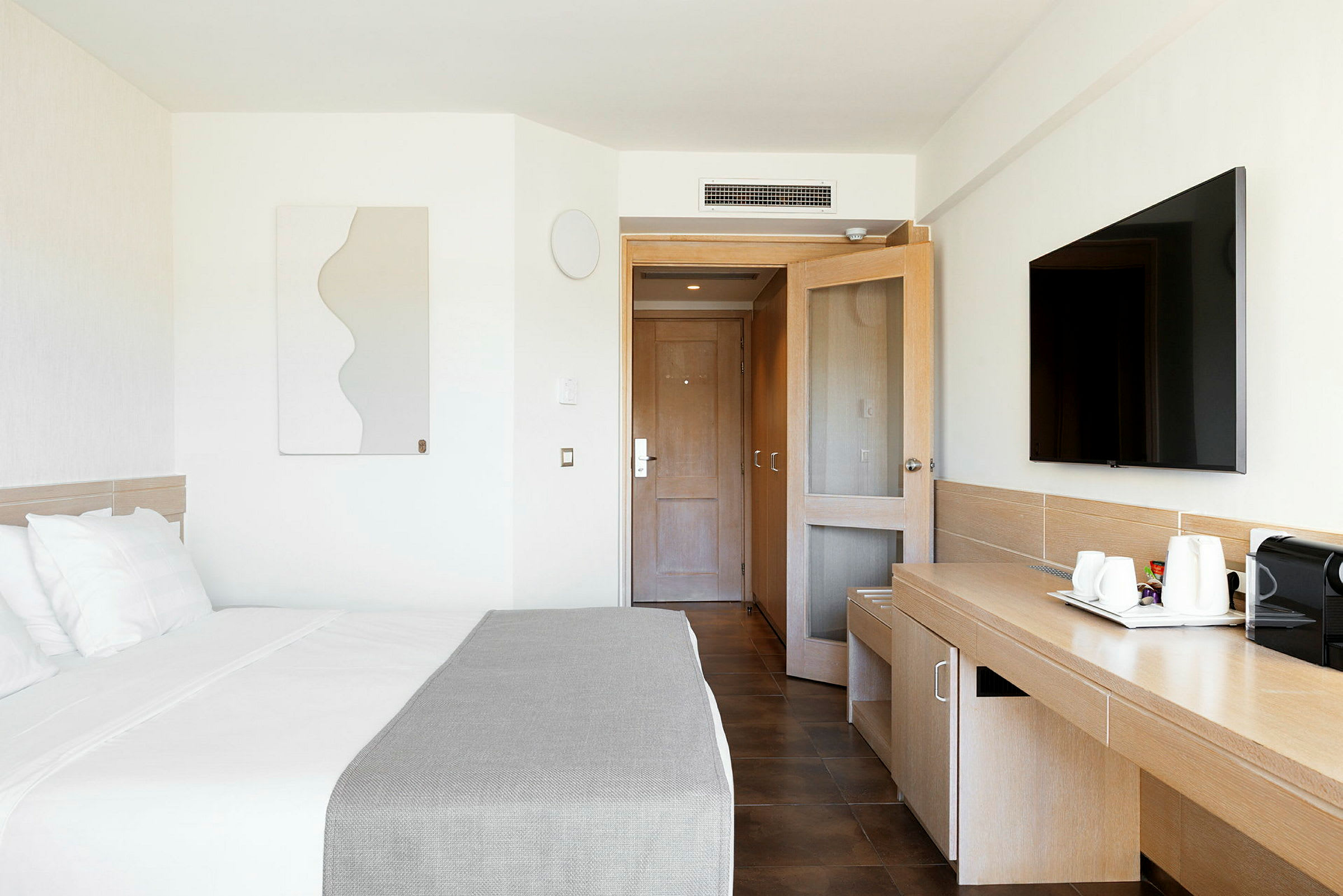 Et moderne hotelværelse med en seng, et skrivebord, et fjernsyn og en dør i baggrunden. (AI-genereret tekst)
