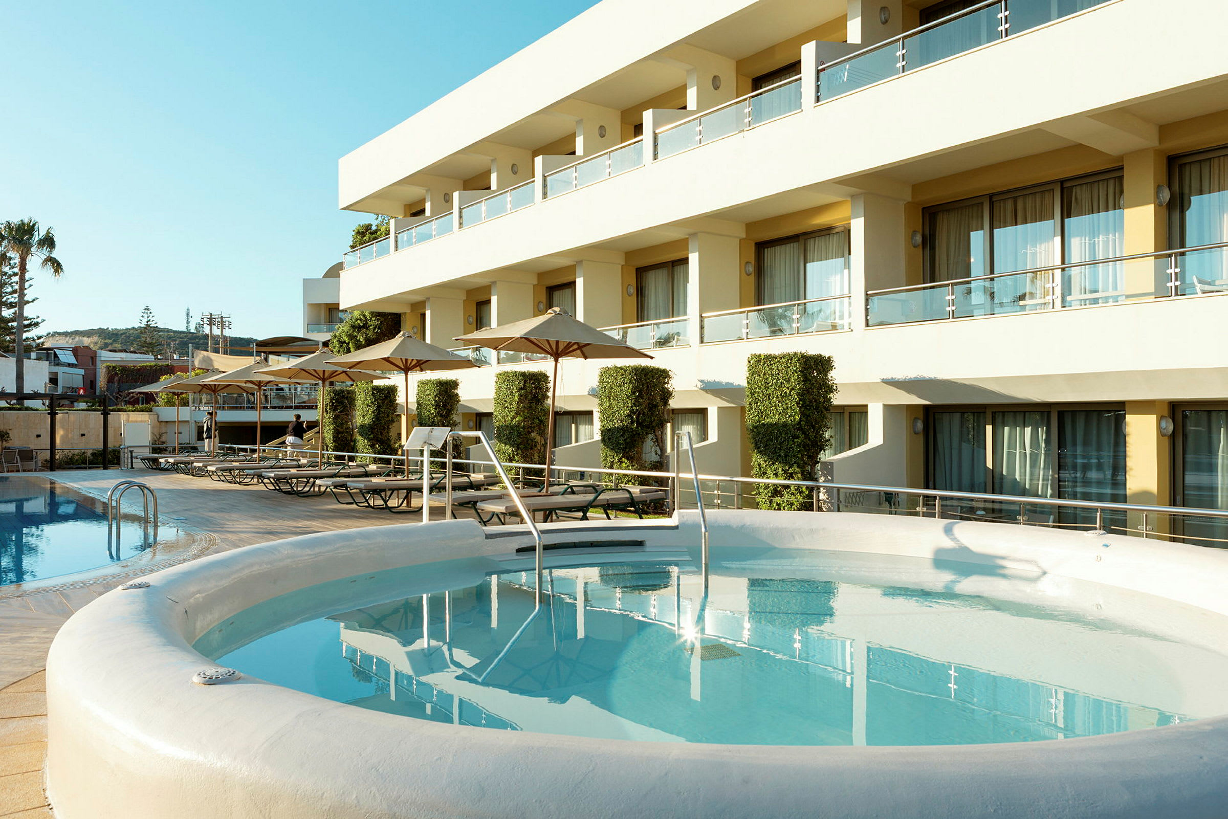 En udendørs pool ved en moderne bygning med glasbalkoner og liggestole på terrassen. (AI-genereret tekst)