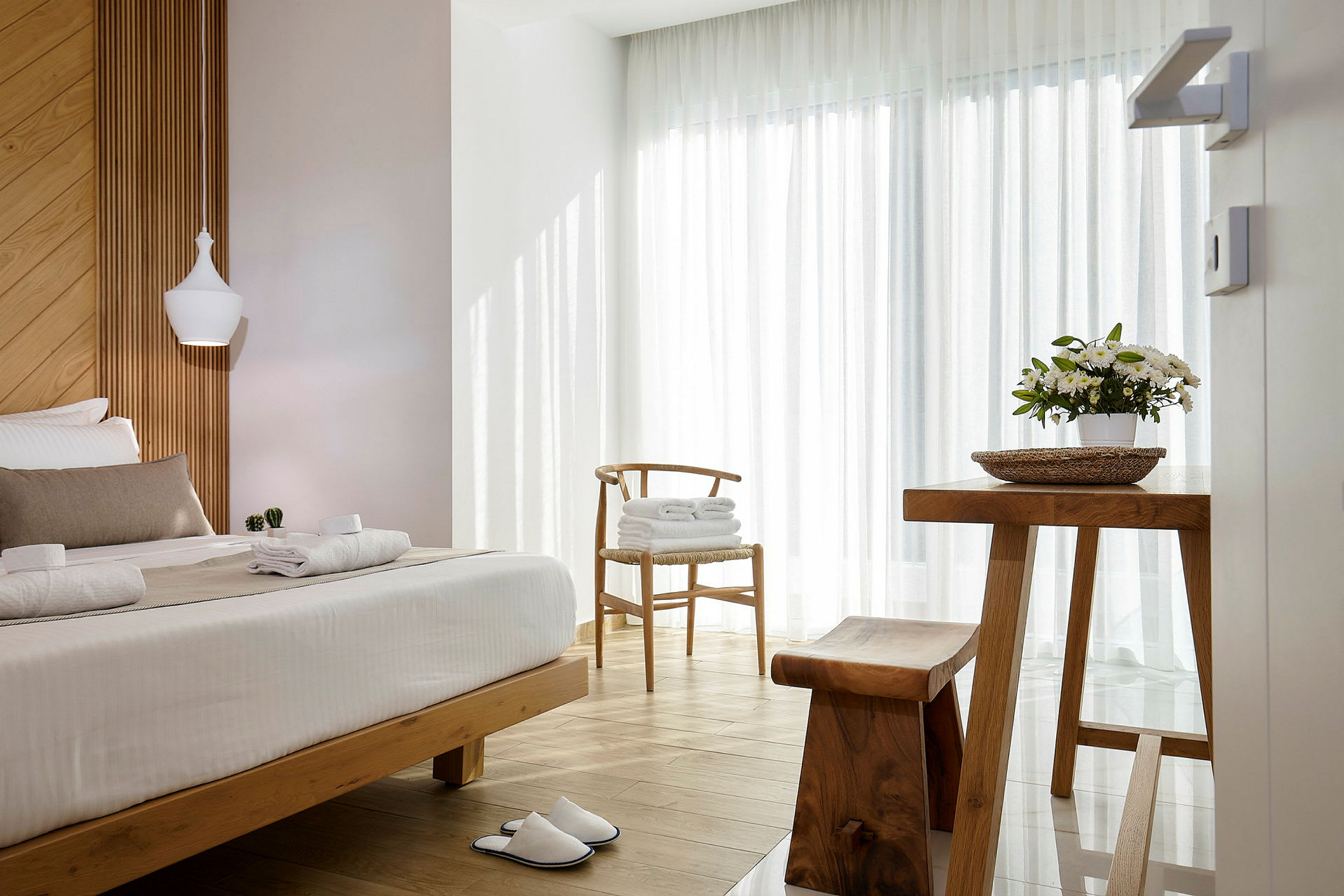 Et lyst og luftigt hotelværelse med trædetaljer, en opredt seng, en blomsterpotte på et bord og et par hvide tøfler på gulvet. (AI-genereret tekst)