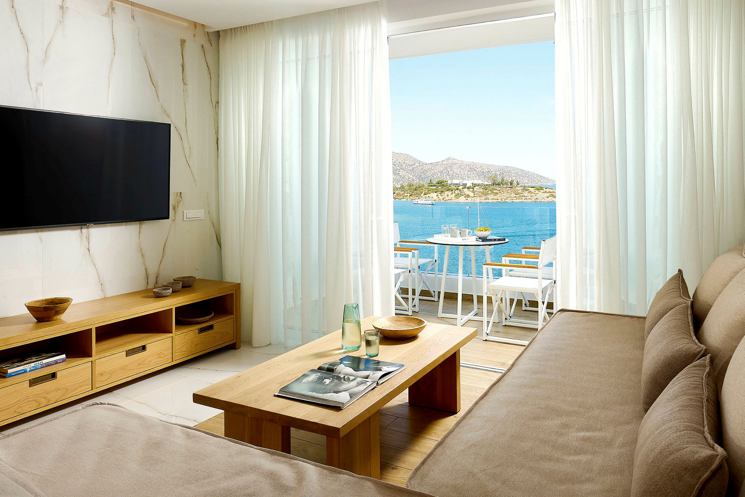 En stue med sofa, sofabord, fjernsyn og udsigt over havet gennem en balkon med siddepladser. (AI-genereret tekst)