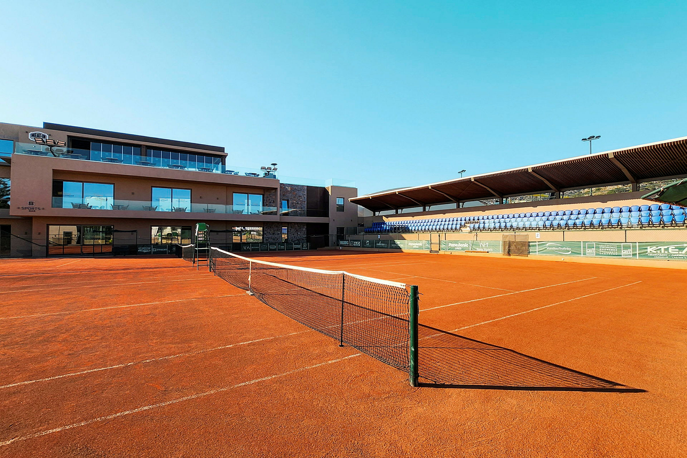 En tennisbane med rødgrus og net foran en bygning og tribuner. (AI-genereret tekst)