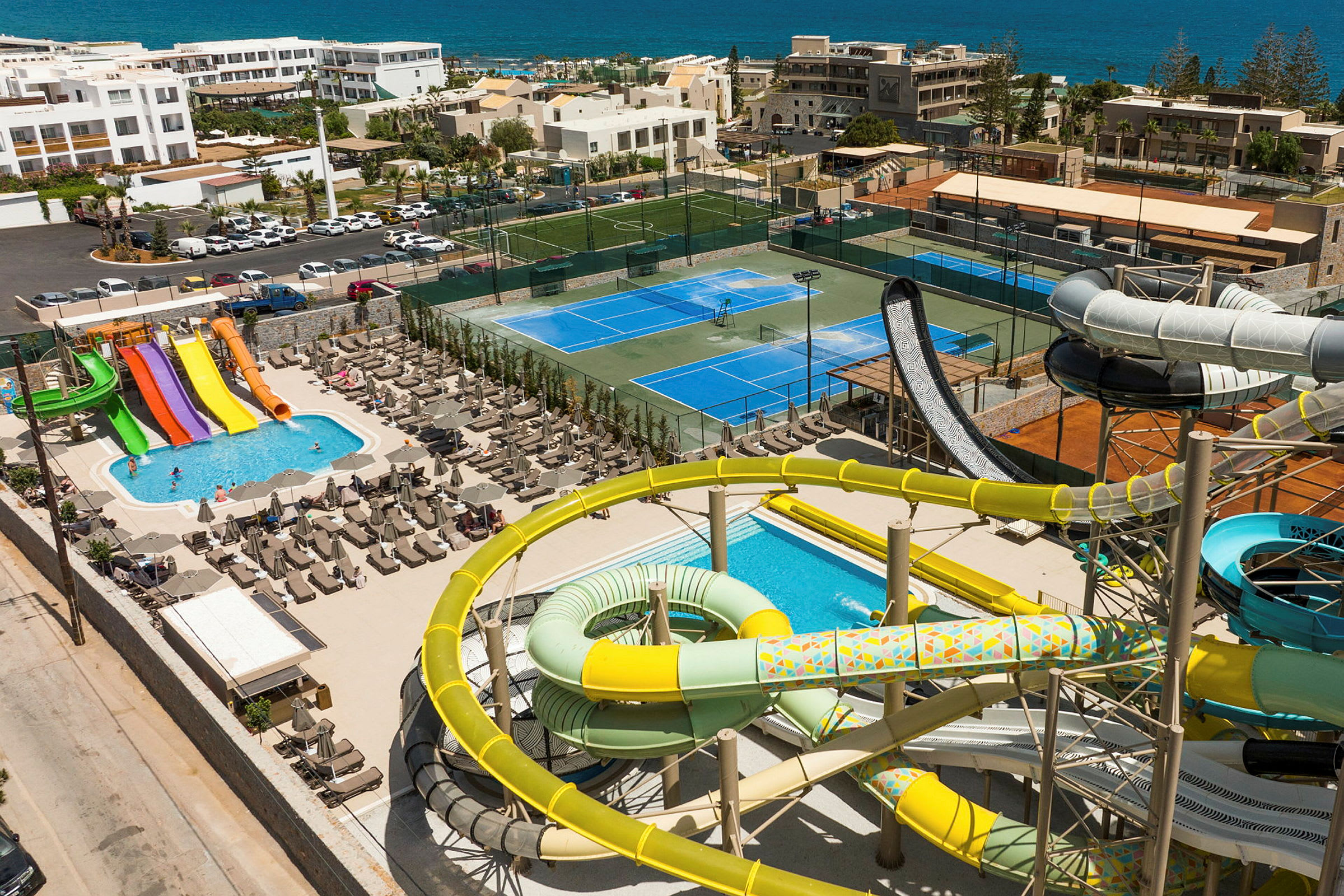 Billede af et feriested med vandrutsjebaner, pool, tennisbaner og udsigt over havet. (AI-genereret tekst)