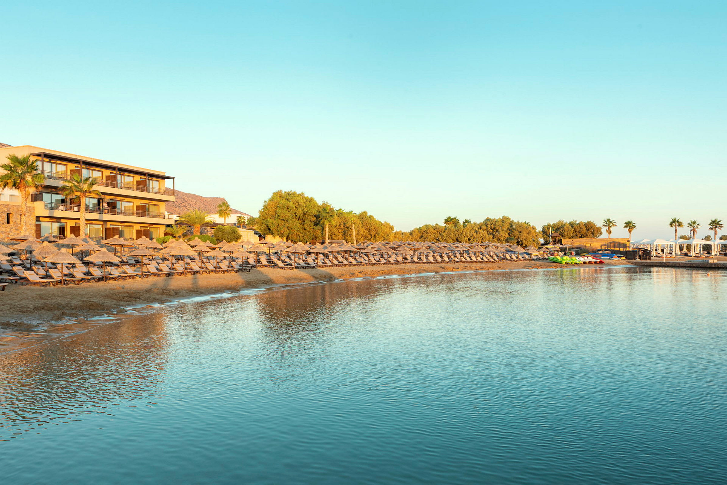 En strand med liggestole og parasoller ved et hotel med udsigt over roligt vand. (AI-genereret tekst)