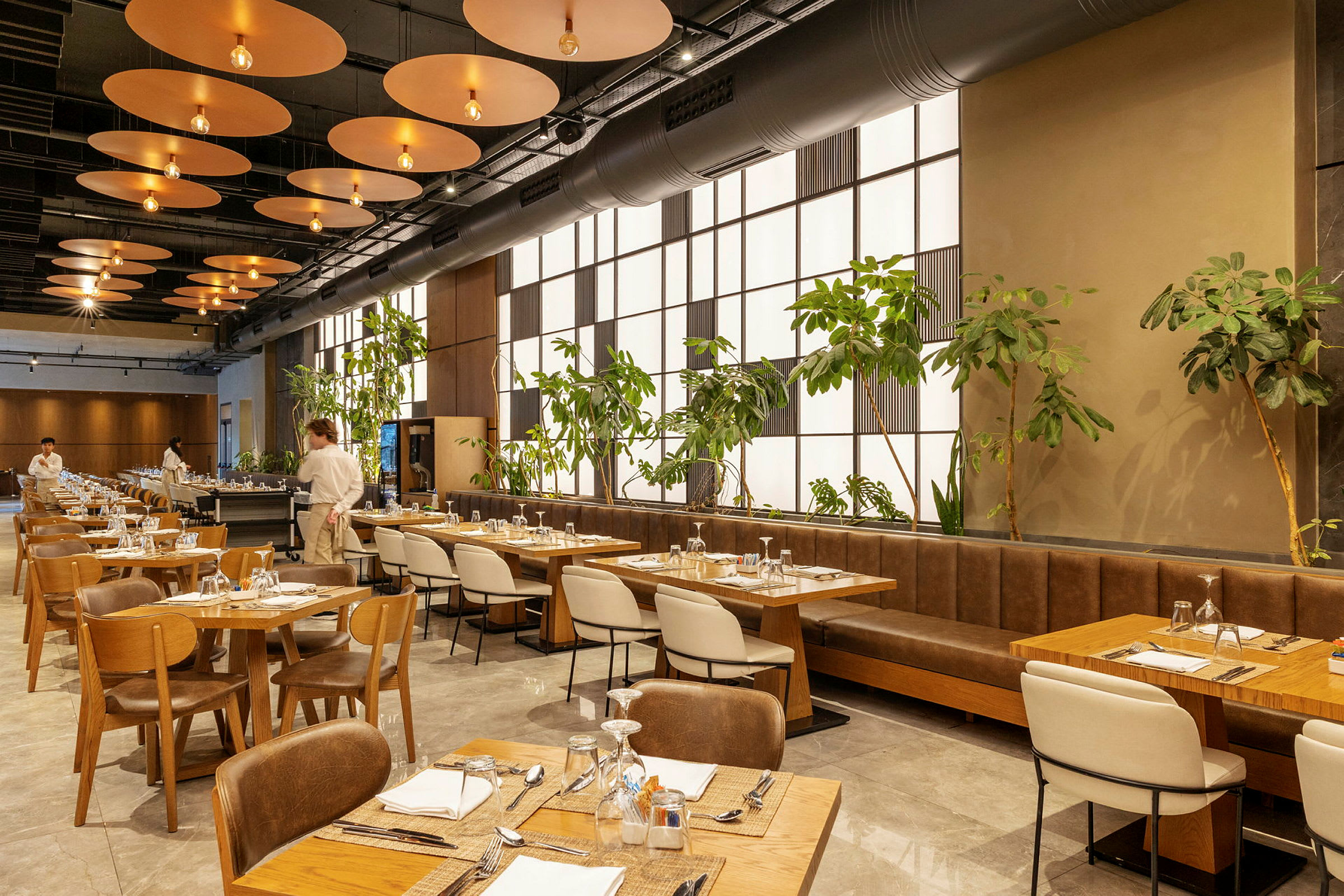 En moderne restaurant med træborde og stole, grønne planter langs en væg og en tjener synlig i baggrunden. (AI-genereret tekst)
