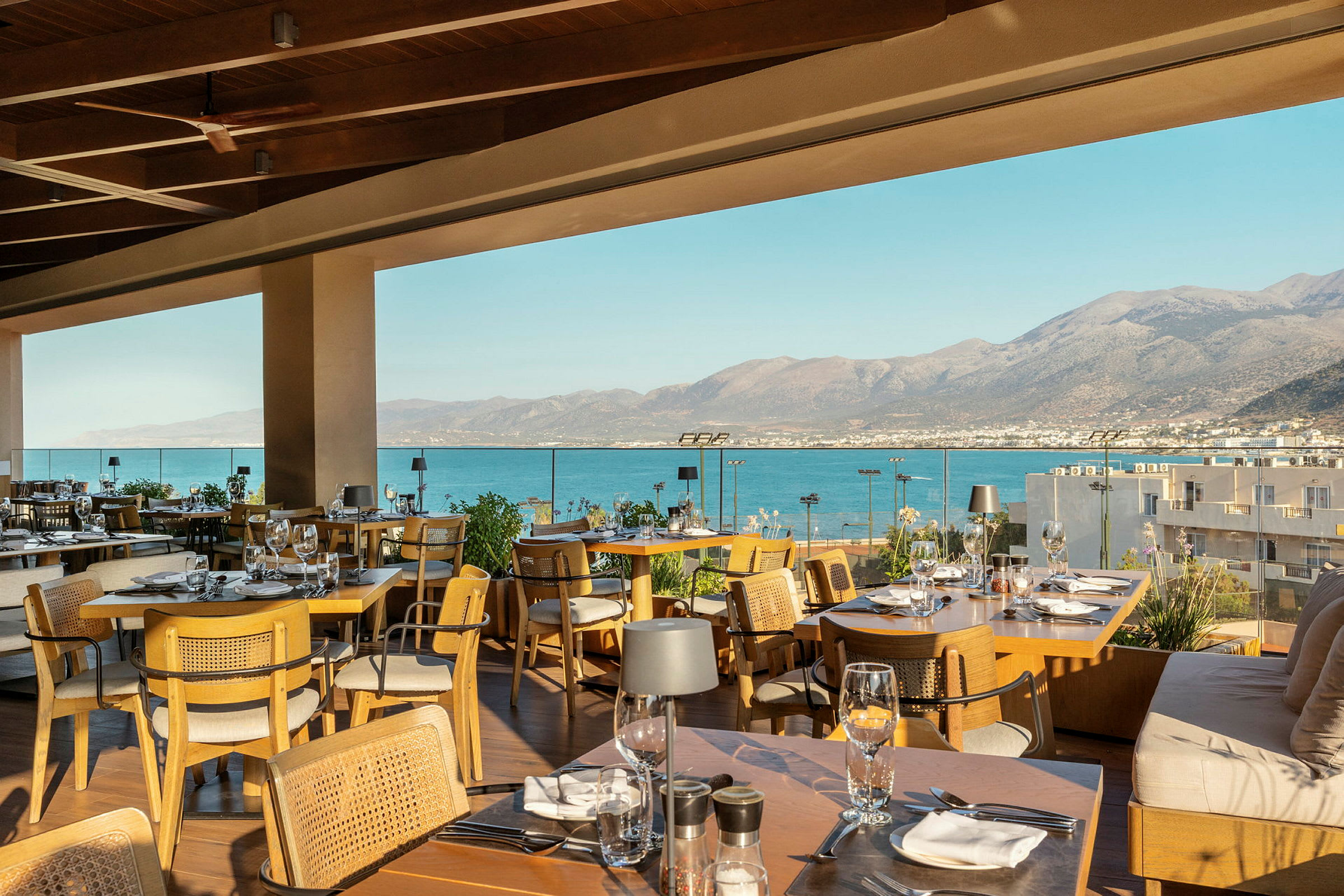 En restaurant med udsigt over havet fra en terrasse. Udendørsområdet er møbleret med træborde og stole, og bjergene i baggrunden strækker sig ud over vandet. (AI-genereret tekst)