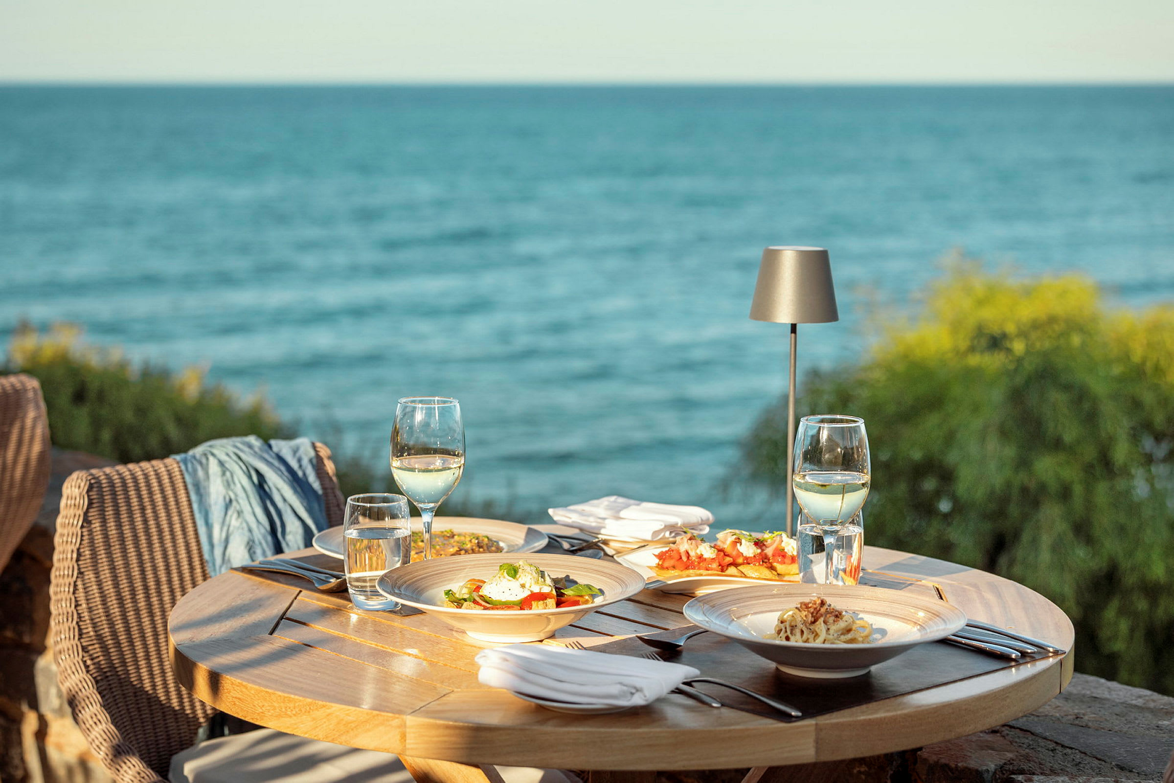 Et udendørs spiseområde ved havet med et rundt bord fyldt med tallerkener med mad, vinglas og bestik. (AI-genereret tekst)