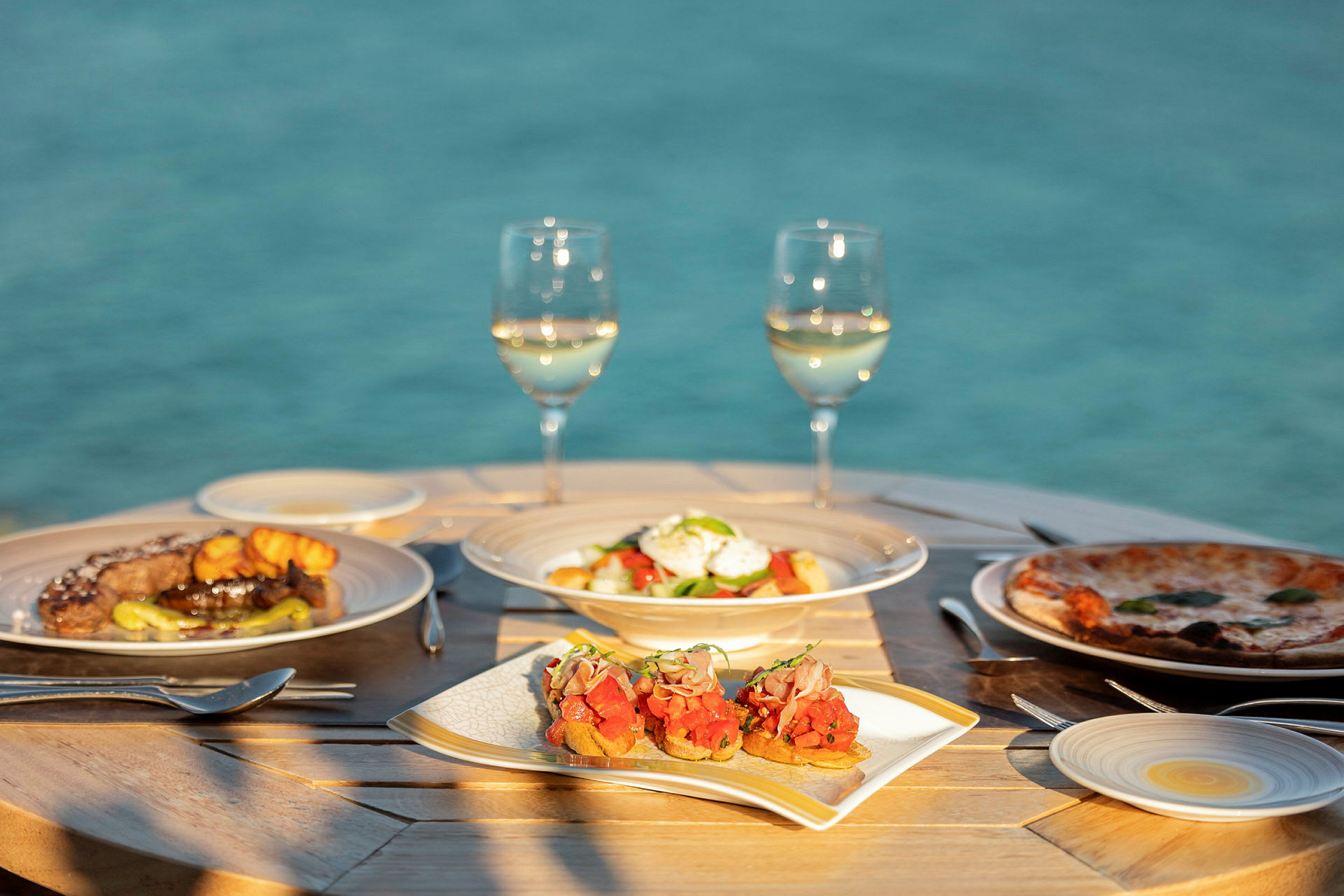 Et bord med et måltid bestående af et udvalg af retter og to glas hvidvin med en baggrund af havet. (AI-genereret tekst)