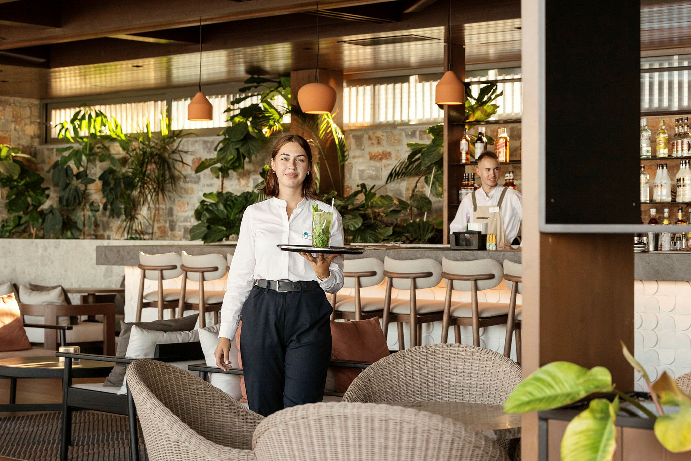 En tjener bærer en bakke med en drink i en hyggelig lounge med planter og en bardisk i baggrunden. (AI-genereret tekst)