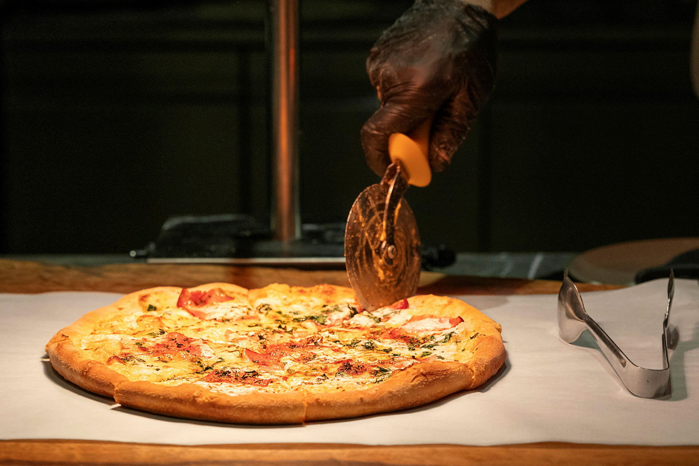 En pizza på et bagepapir bliver skåret med en pizzaskærer holdt af en hånd i sort handske. (AI-genereret tekst)