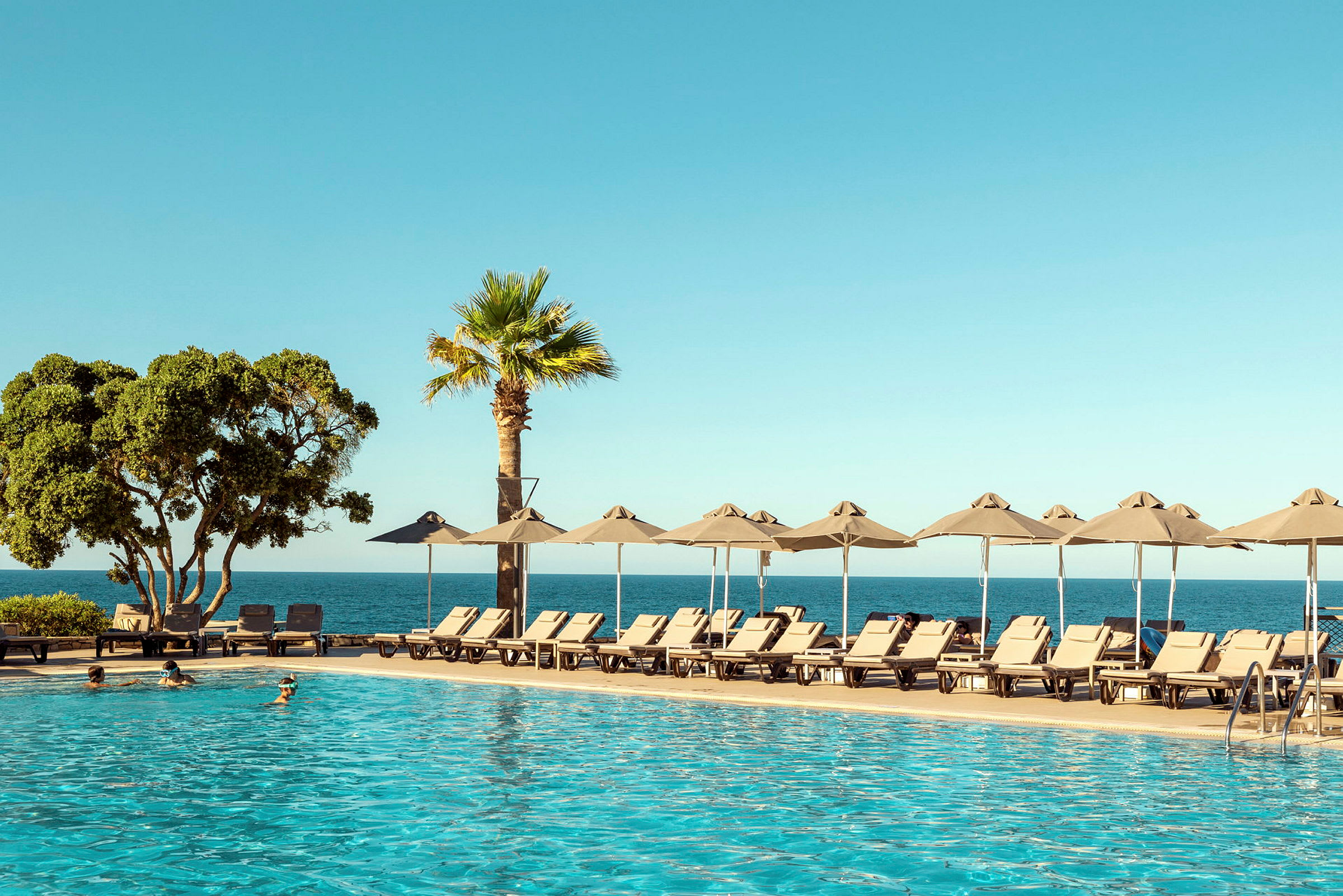 En pool i forgrunden med solsenge og parasoller ved siden. Bagved ses havet og en klar blå himmel. (AI-genereret tekst)