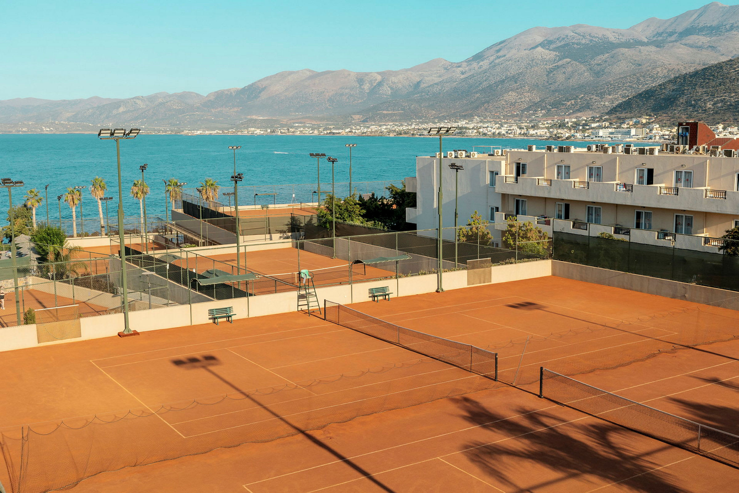 Et billede af flere tennisbaner lavet af grus, beliggende ved kysten tæt på havet med bjerge i baggrunden. (AI-genereret tekst)