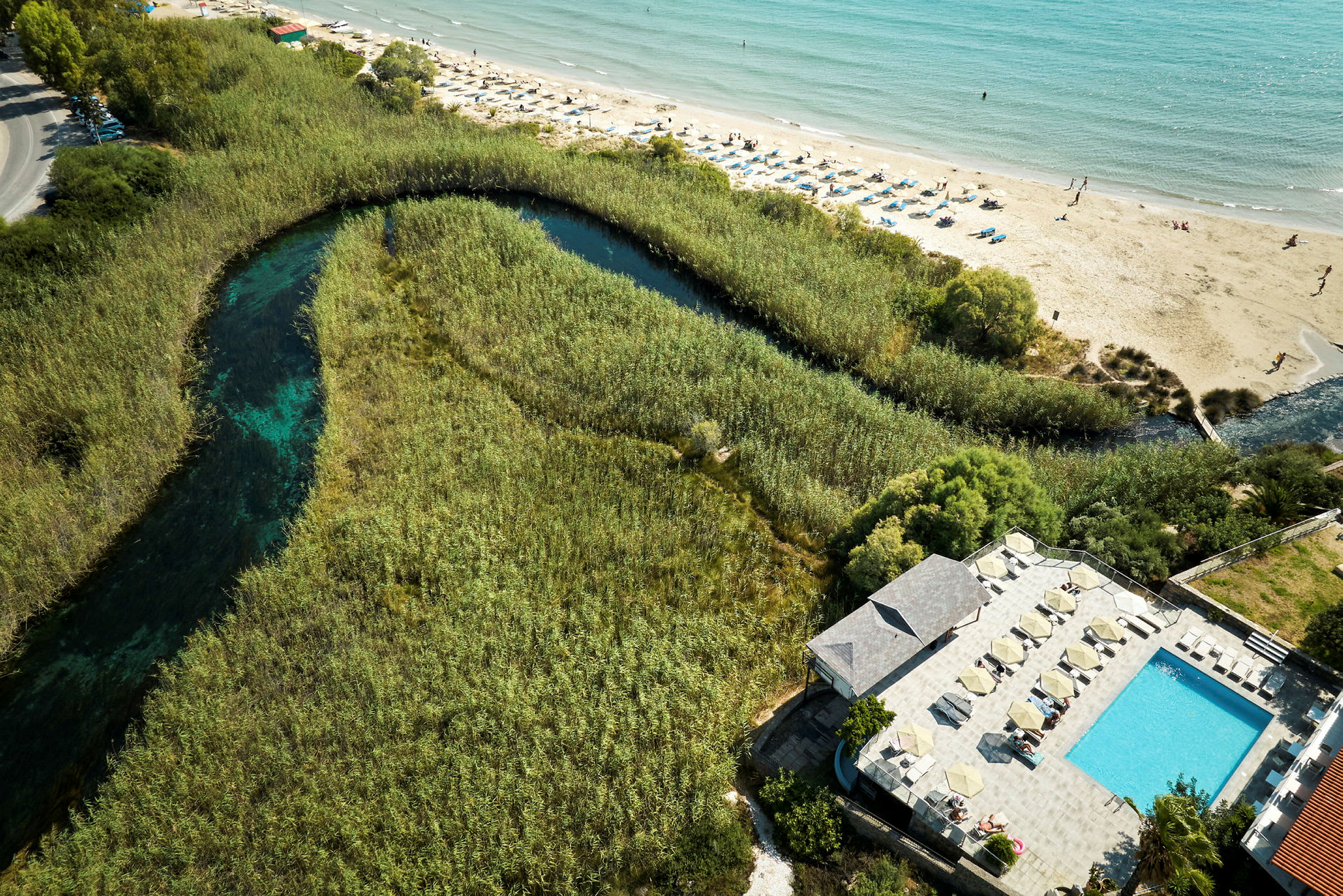 Luftfoto af et landskab med en pool, en strand med liggestole og et vandløb omgivet af vegetation. (AI-genereret tekst)