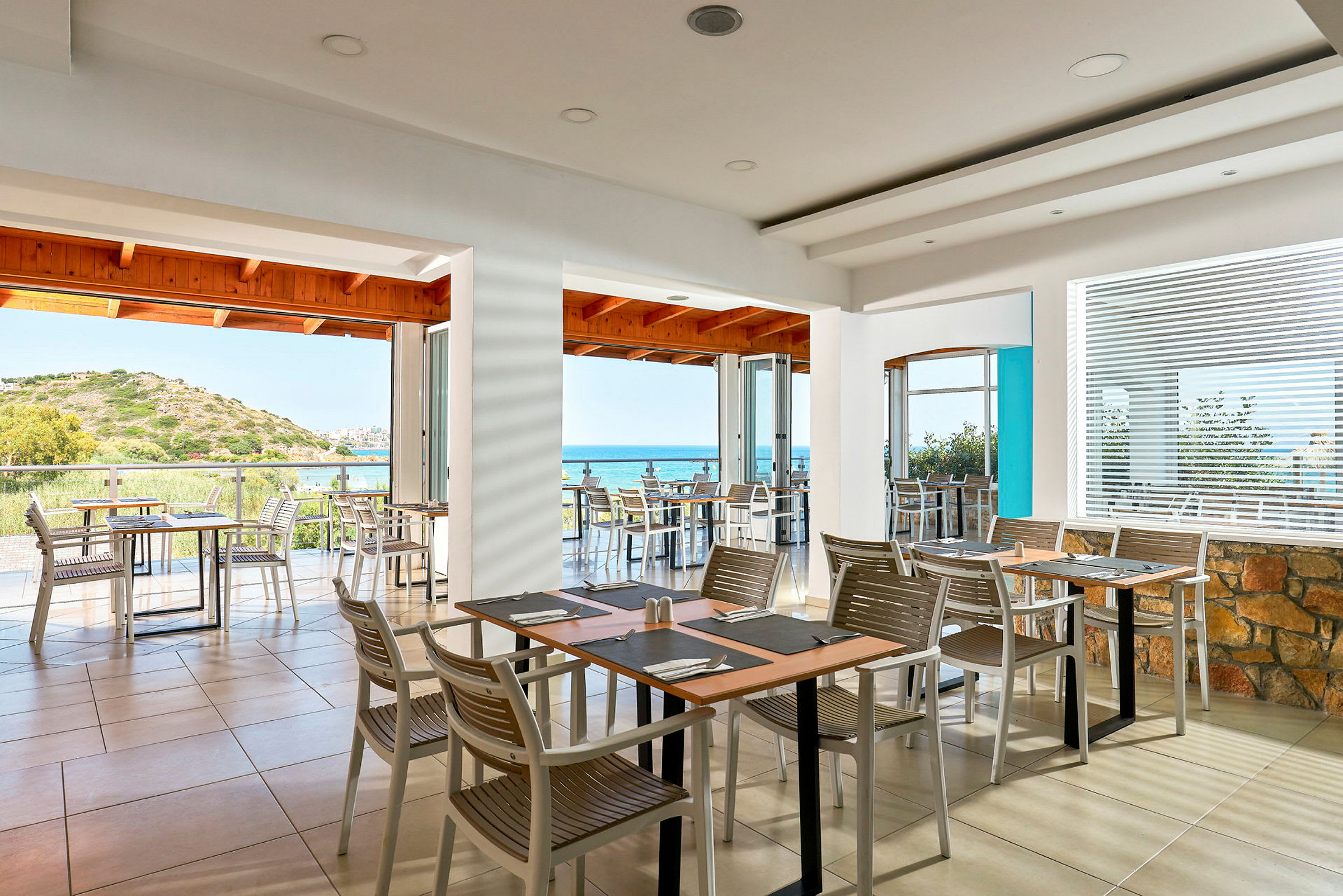 En restaurant med udsigt over havet, med borde og stole indendørs og på en terrasse. (AI-genereret tekst)