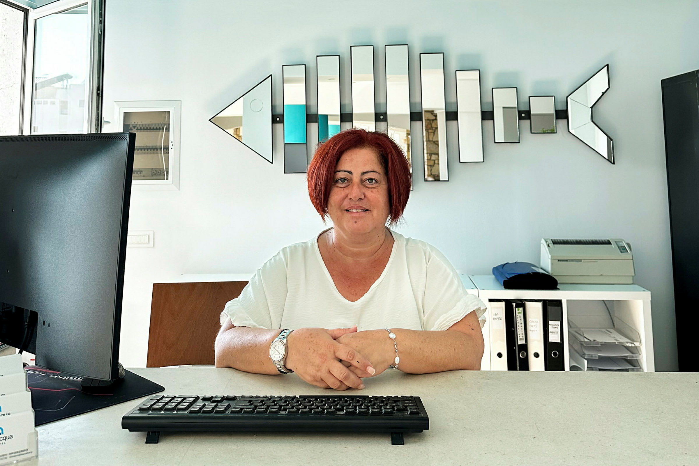 Person sidder ved et skrivebord med tastatur, computerskærm og hylder i baggrunden. (AI-genereret tekst)