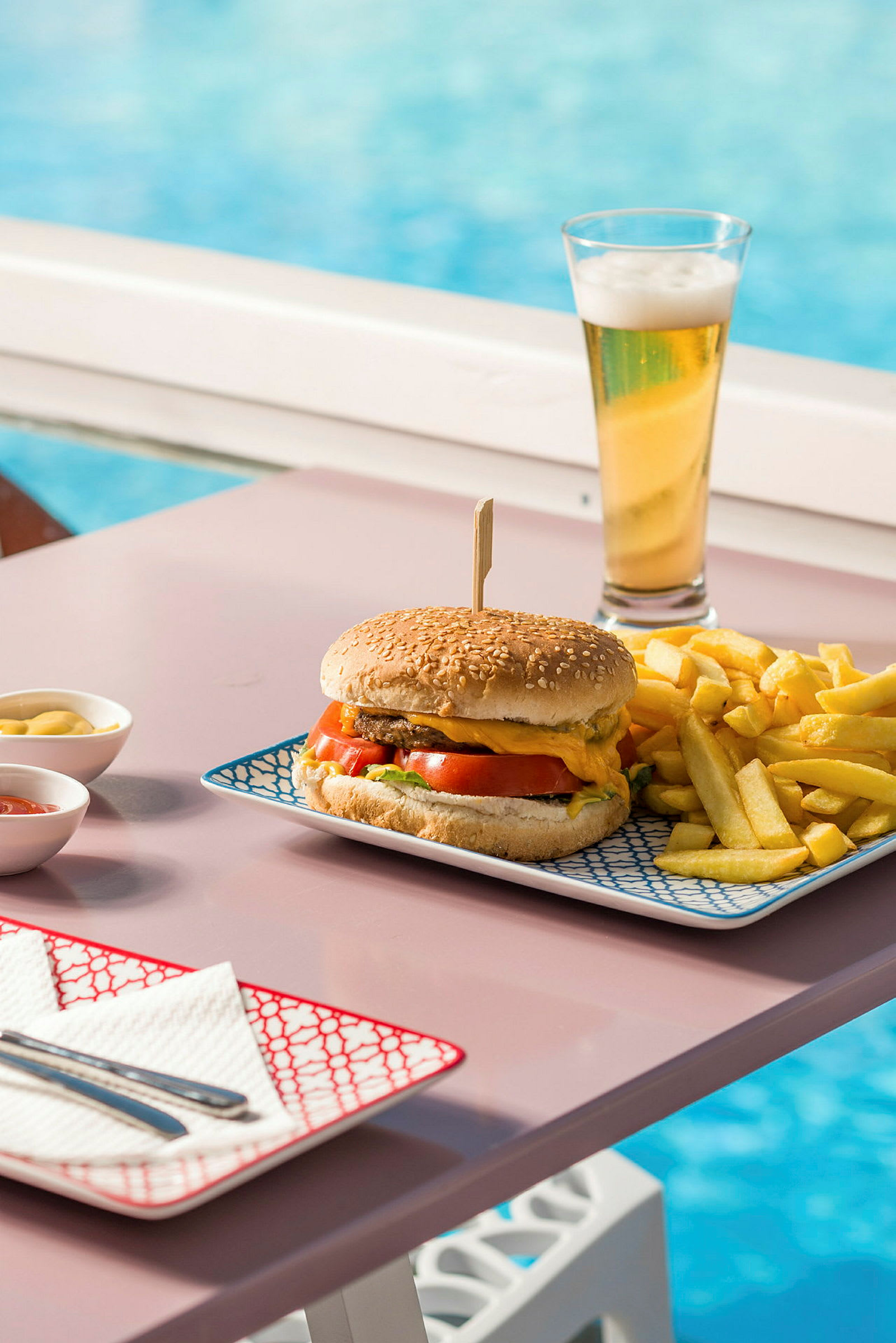 En tallerken med burger, pommes frites og en øl tæt på en pool. (AI-genereret tekst)