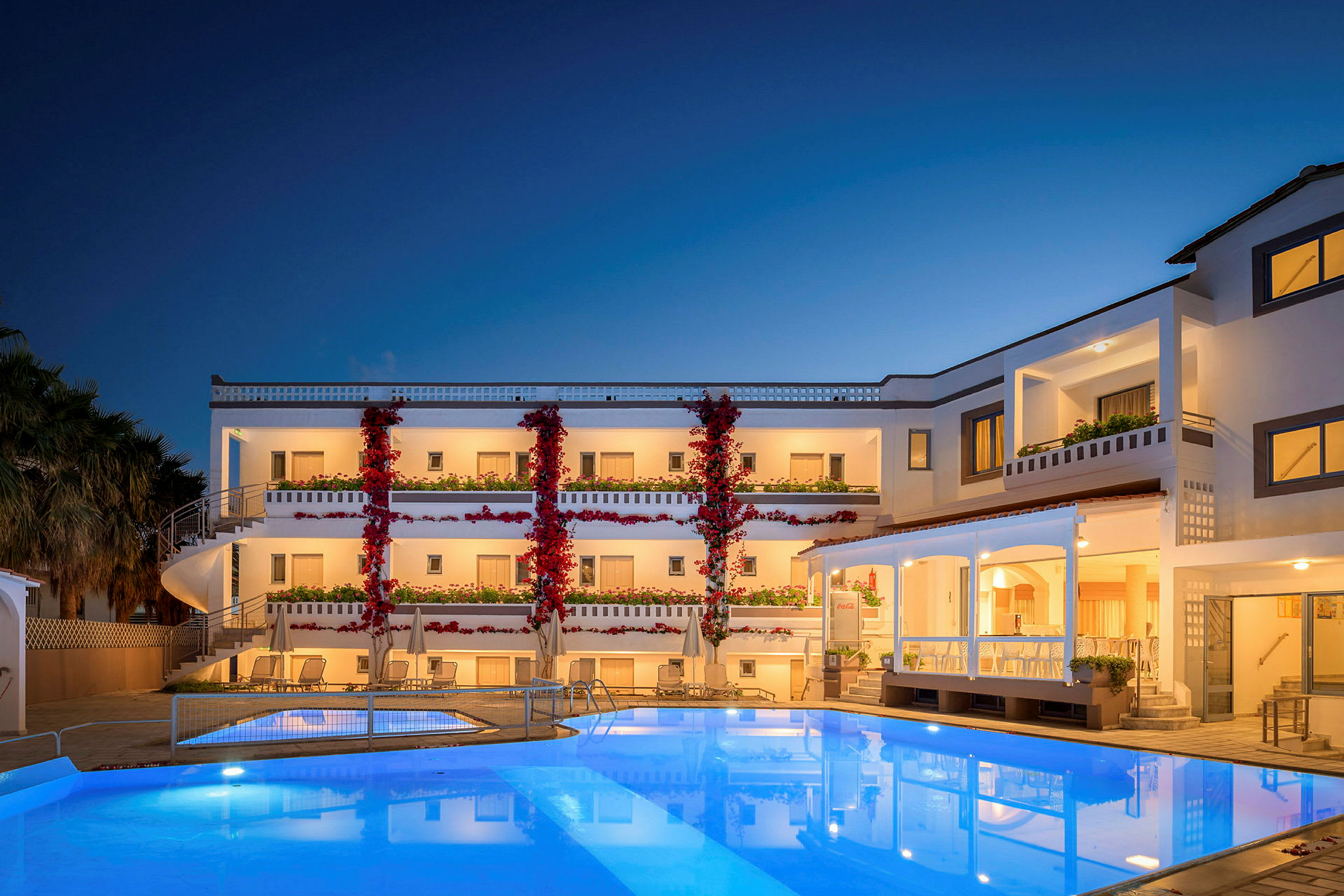 Et hotel med tre etager, med hvid facade og røde blomster, beliggende ved en oplyst pool om aftenen. (AI-genereret tekst)