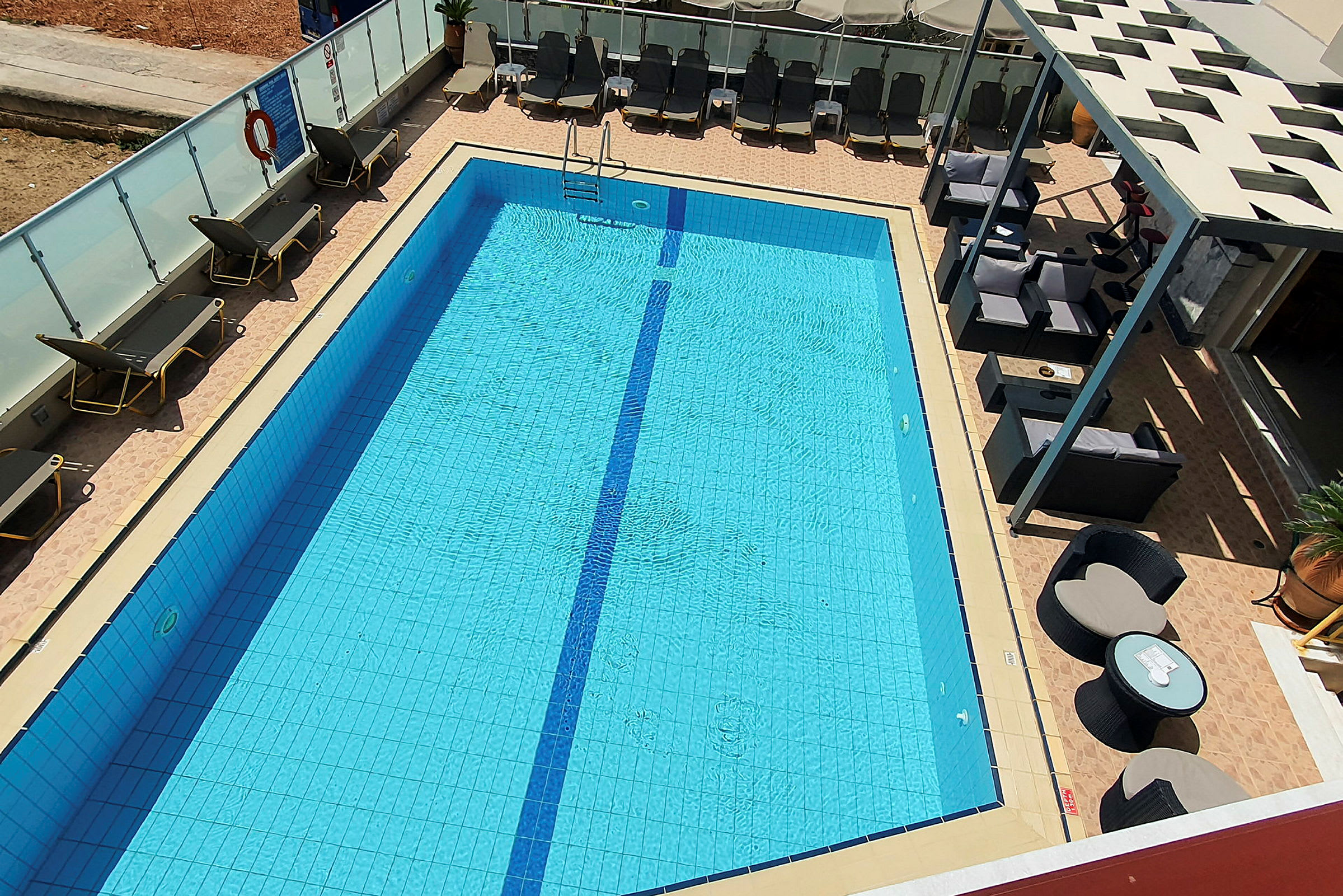 En udendørs swimmingpool med liggestole og møbler rundt om. Poolen er blå med en smal linje langs midten. (AI-genereret tekst)