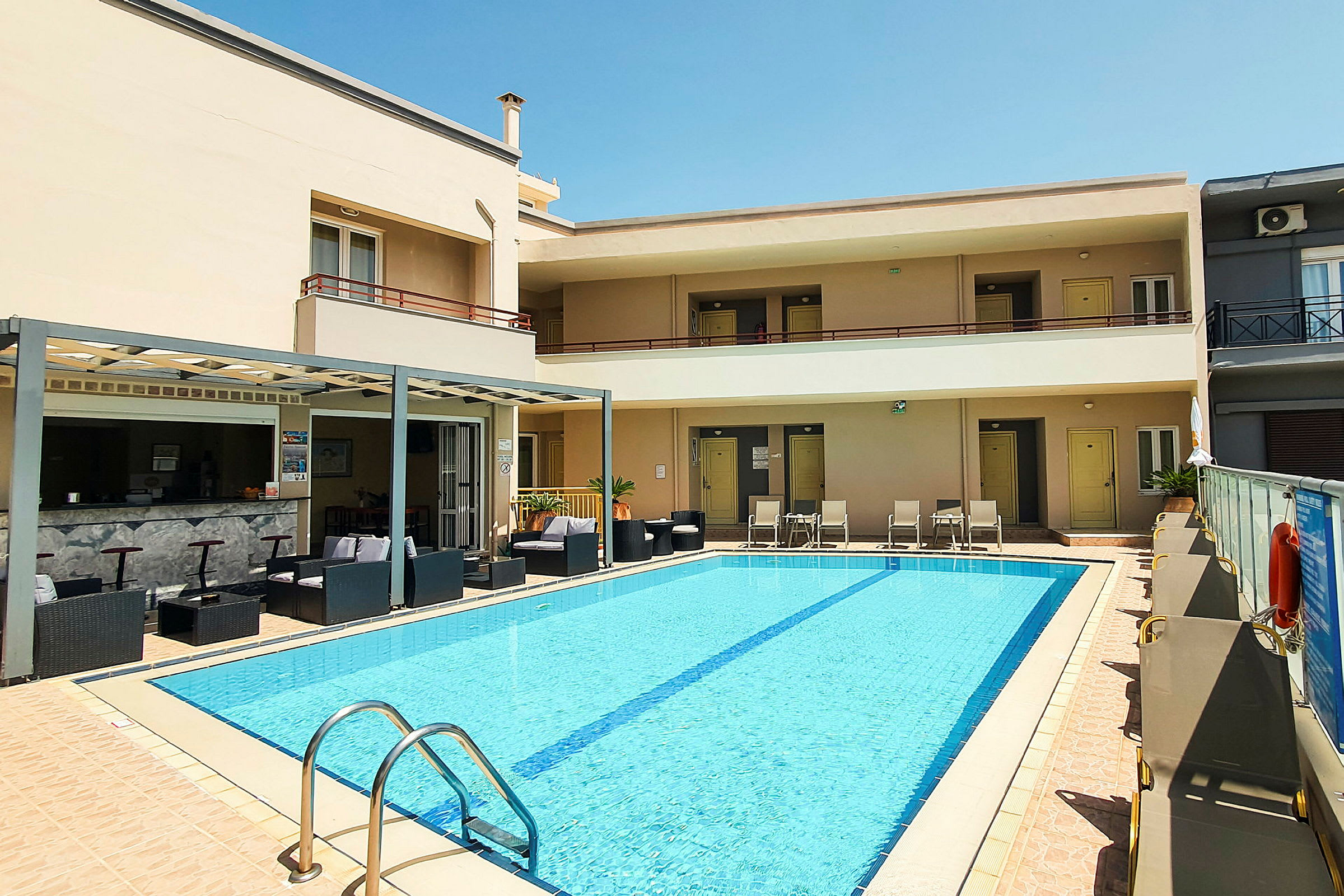 Billede af et hotel med en pool omgivet af liggestole. En bar og siddeområde ses i baggrunden. (AI-genereret tekst)
