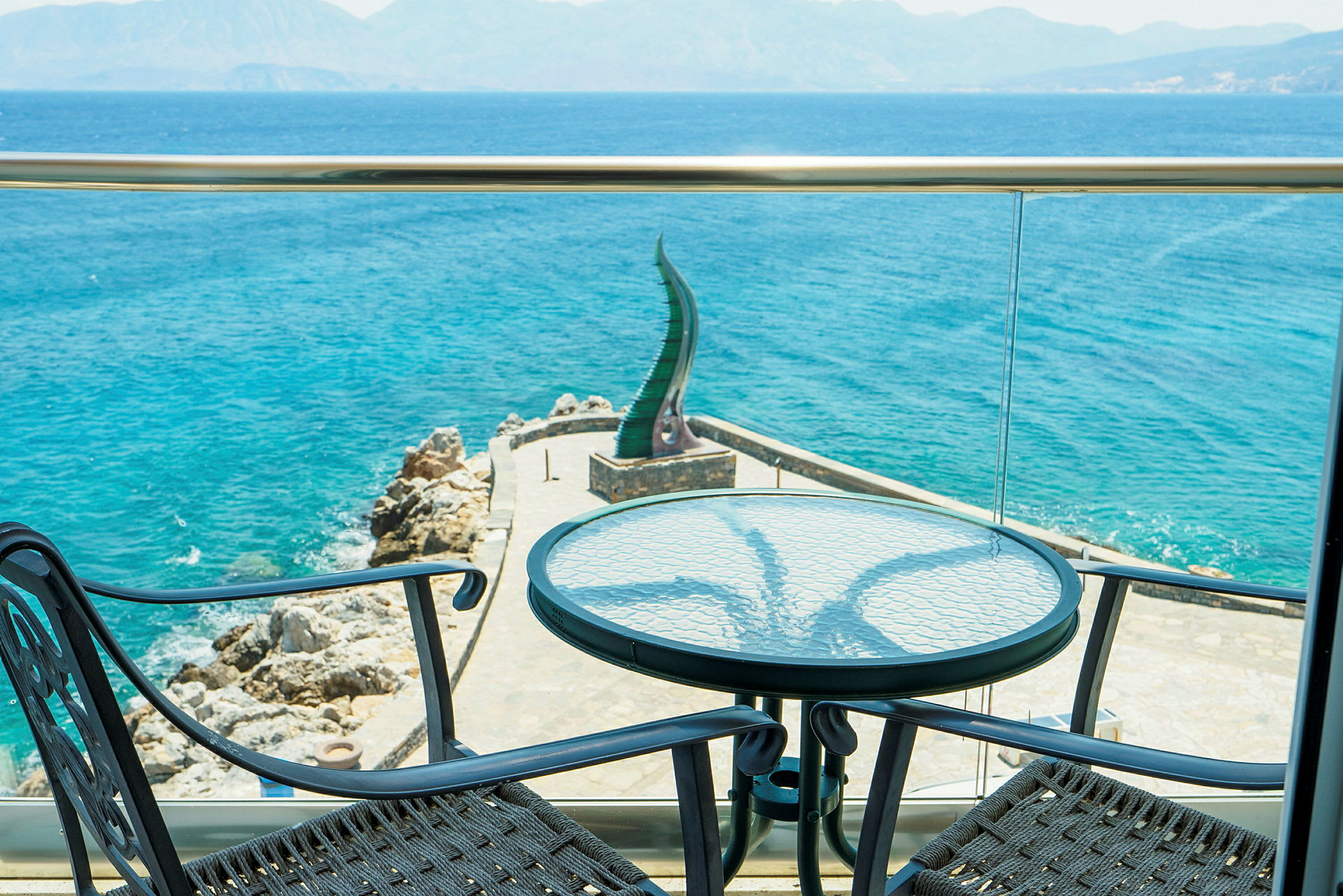 Udsigt fra en terrasse mod havet med to stole og et bord i forgrunden. (AI-genereret tekst)