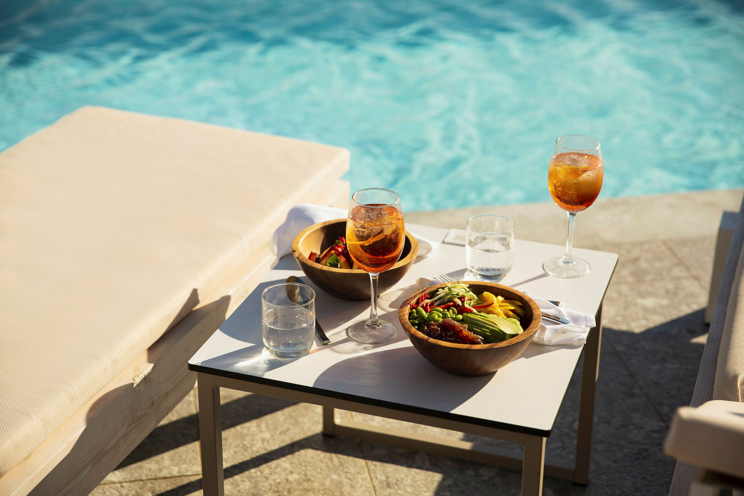 To salatskåle og glas med drikke på et bord ved siden af en pool. (AI-genereret tekst)