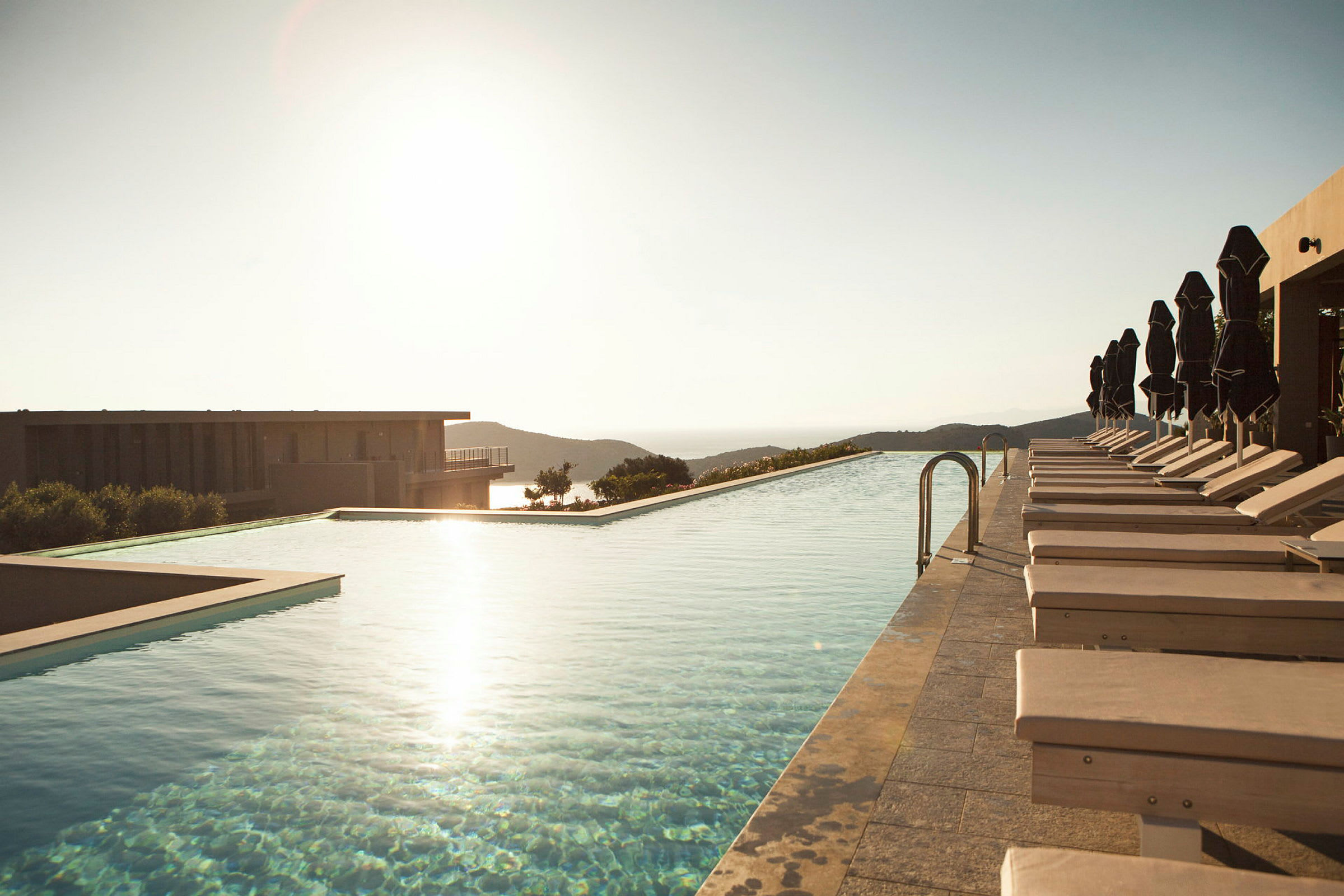 En infinitypool med liggestole og parasoller ved siden af, med udsigt over bakker og hav ved solnedgang. (AI-genereret tekst)