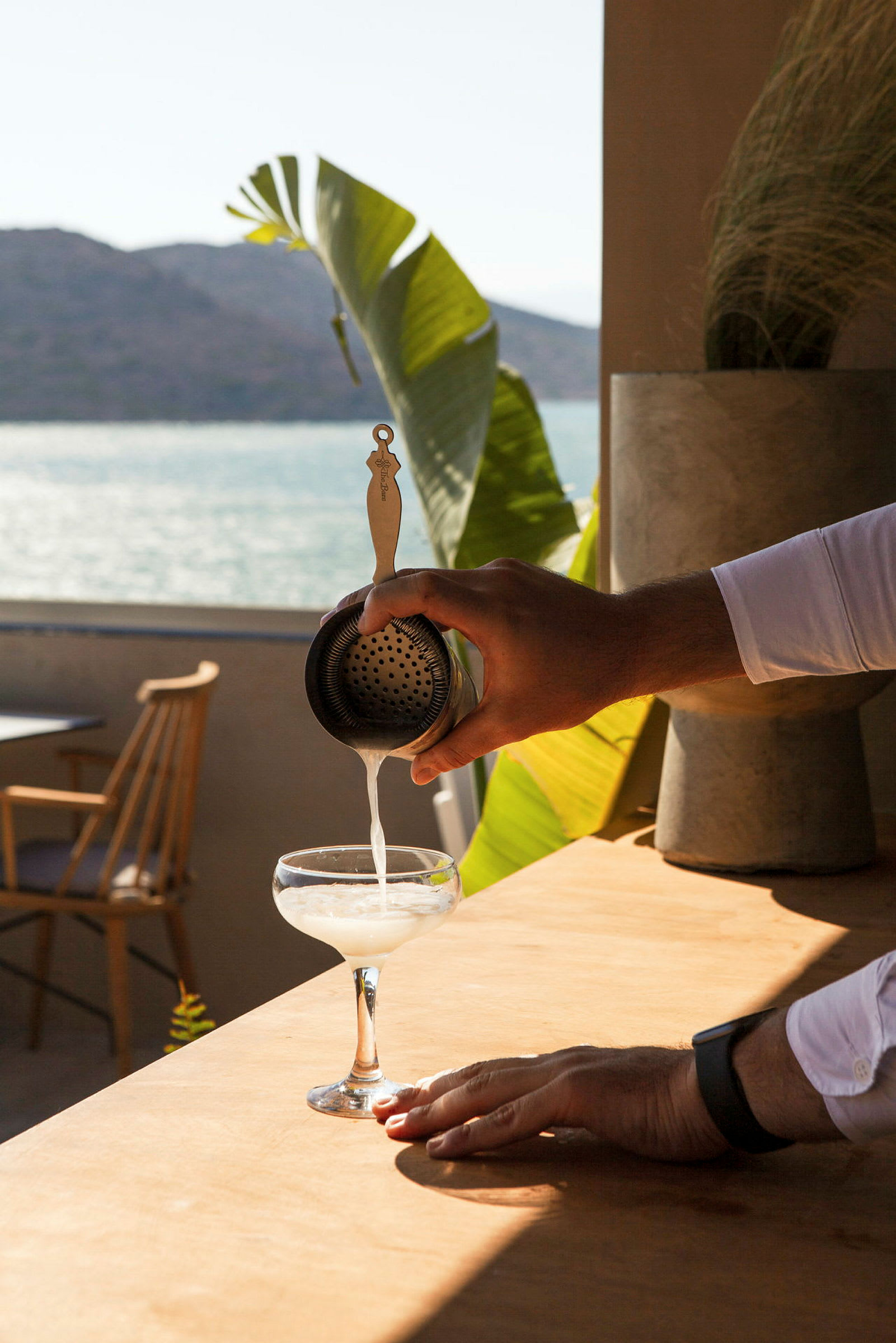 En hånd hælder en drik gennem en si i et cocktailglas, med havudsigt og planter i baggrunden. (AI-genereret tekst)